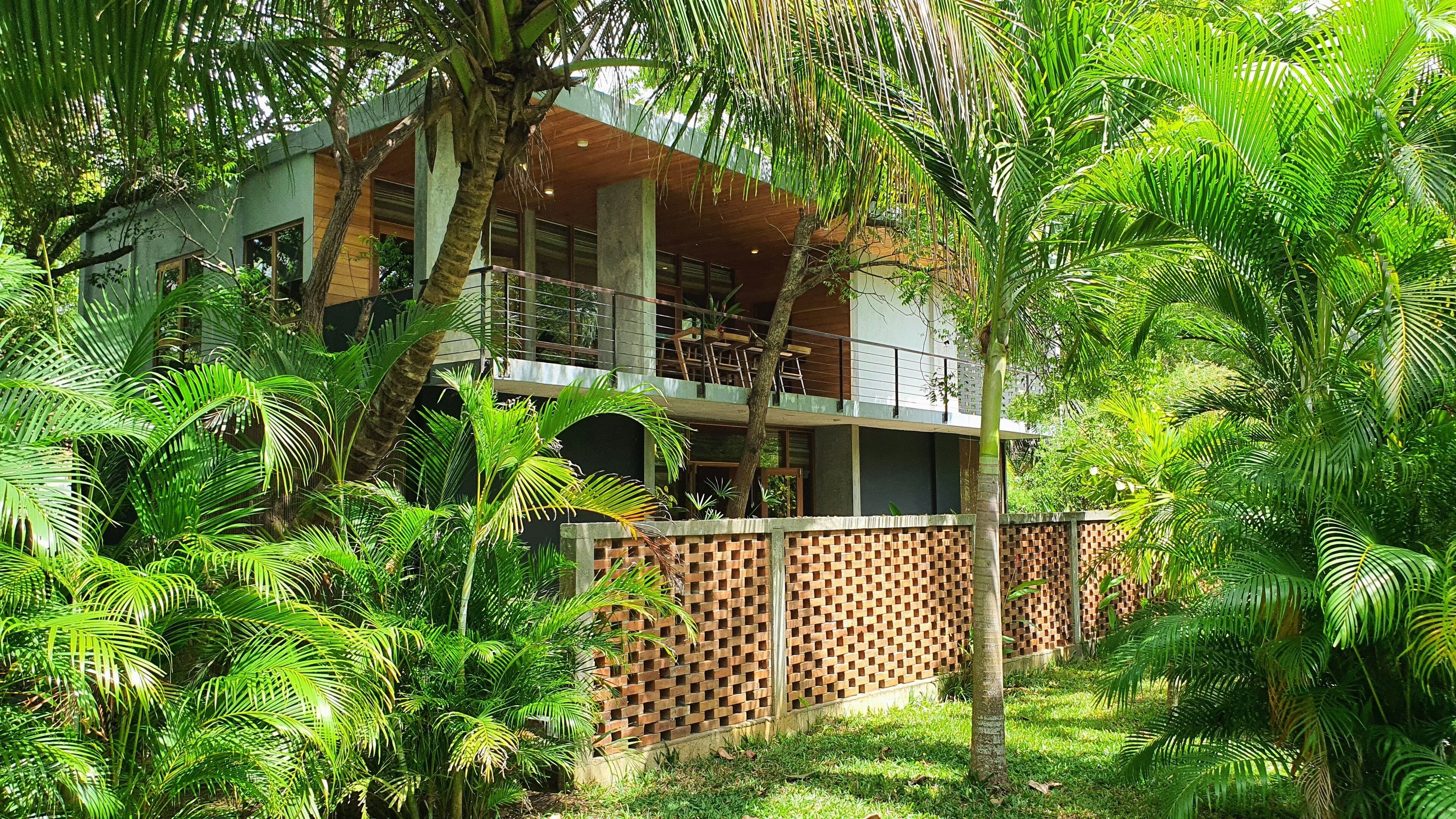 A modern two-story building with a balcony, surrounded by lush greenery and a brick wall.
