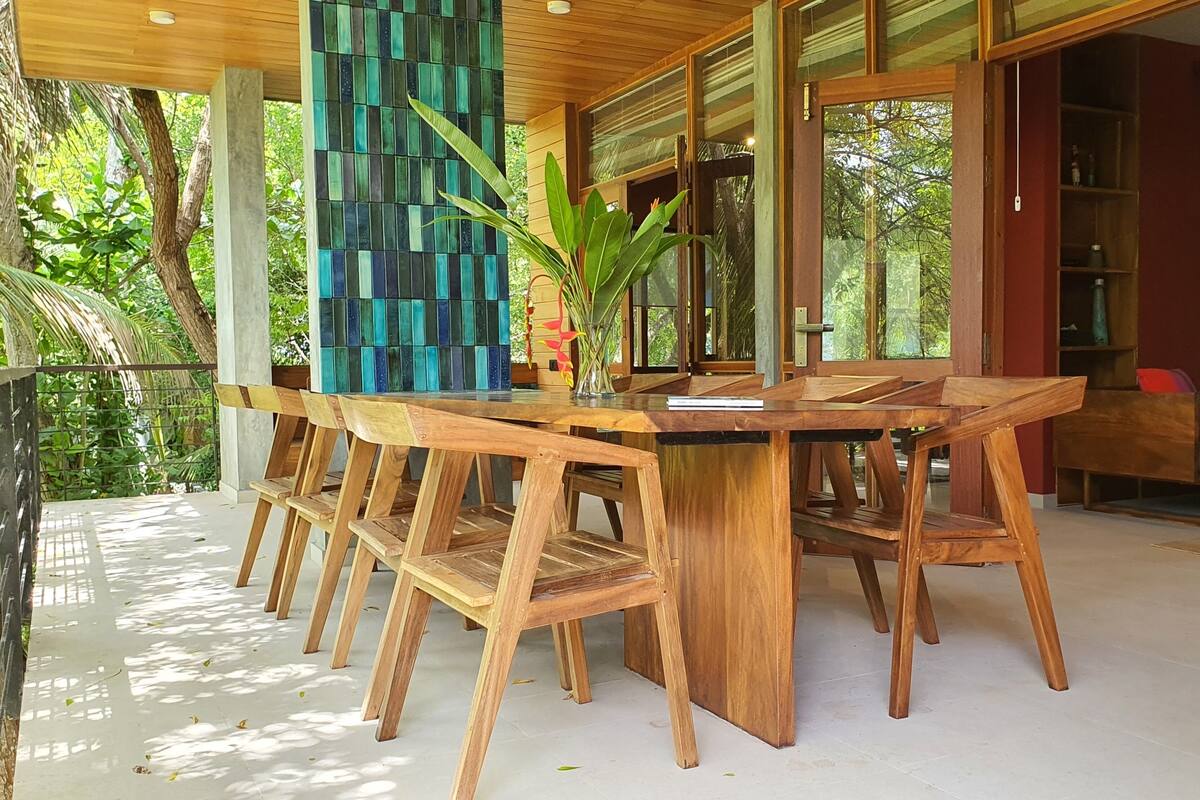 A wooden dining table with chairs, surrounded by glass doors and windows, with a view of greenery outside.