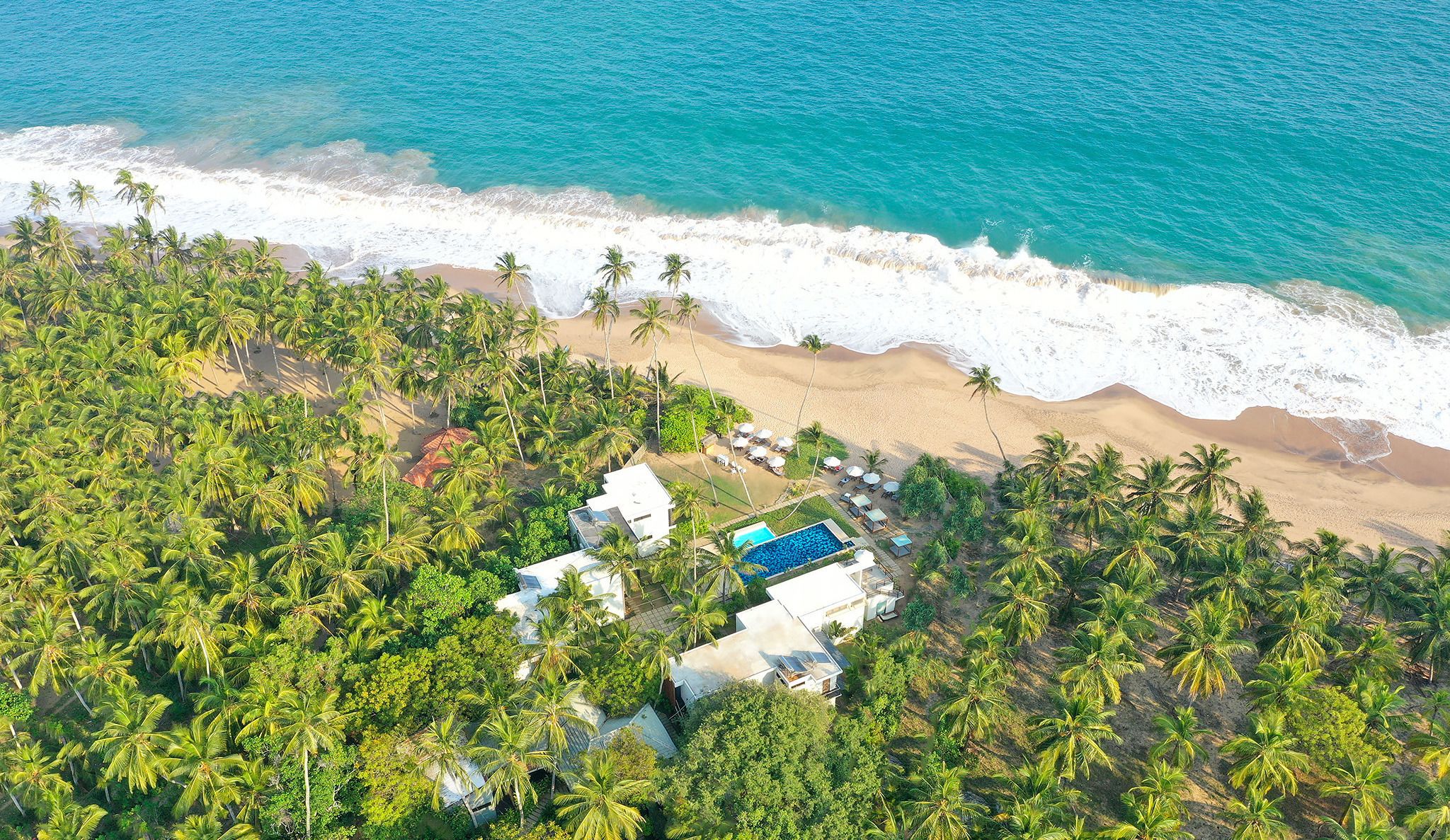 An aerial view of a beach resort with a swimming pool, palm trees, and a sandy beach.