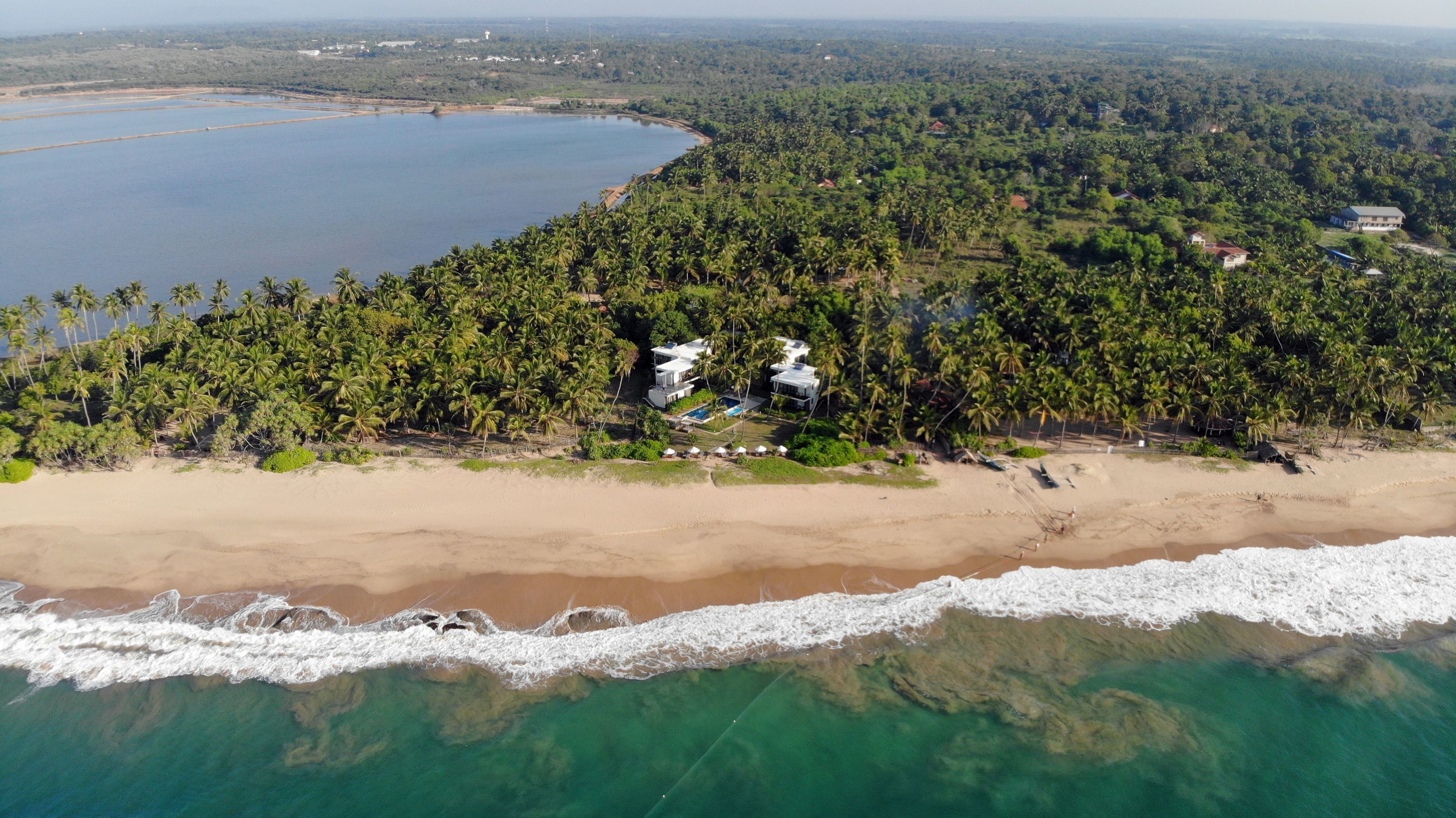 Vista aerea di una zona costiera con spiaggia, palme rigogliose e un piccolo gruppo di edifici con piscina.