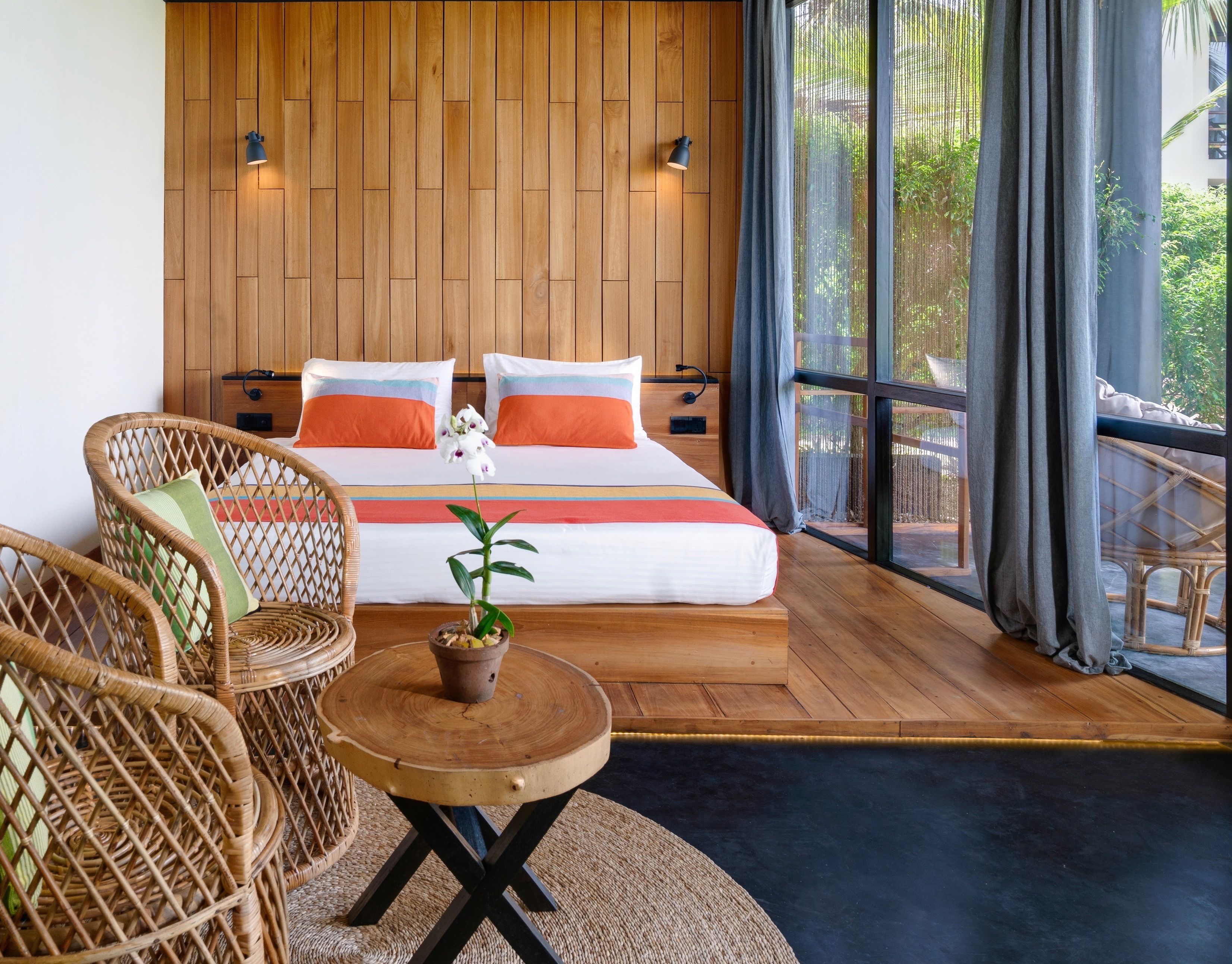 Een slaapkamer met een houten hoofdbord, een bed met oranje en witte beddengoed, twee rieten stoelen, een klein rond tafeltje met een bloempot, en een groot raam met gordijnen.