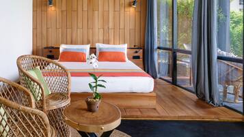 Een slaapkamer met een houten hoofdbord, een bed met oranje en witte beddengoed, twee rieten stoelen, een klein rond tafeltje met een bloempot, en een groot raam met gordijnen.