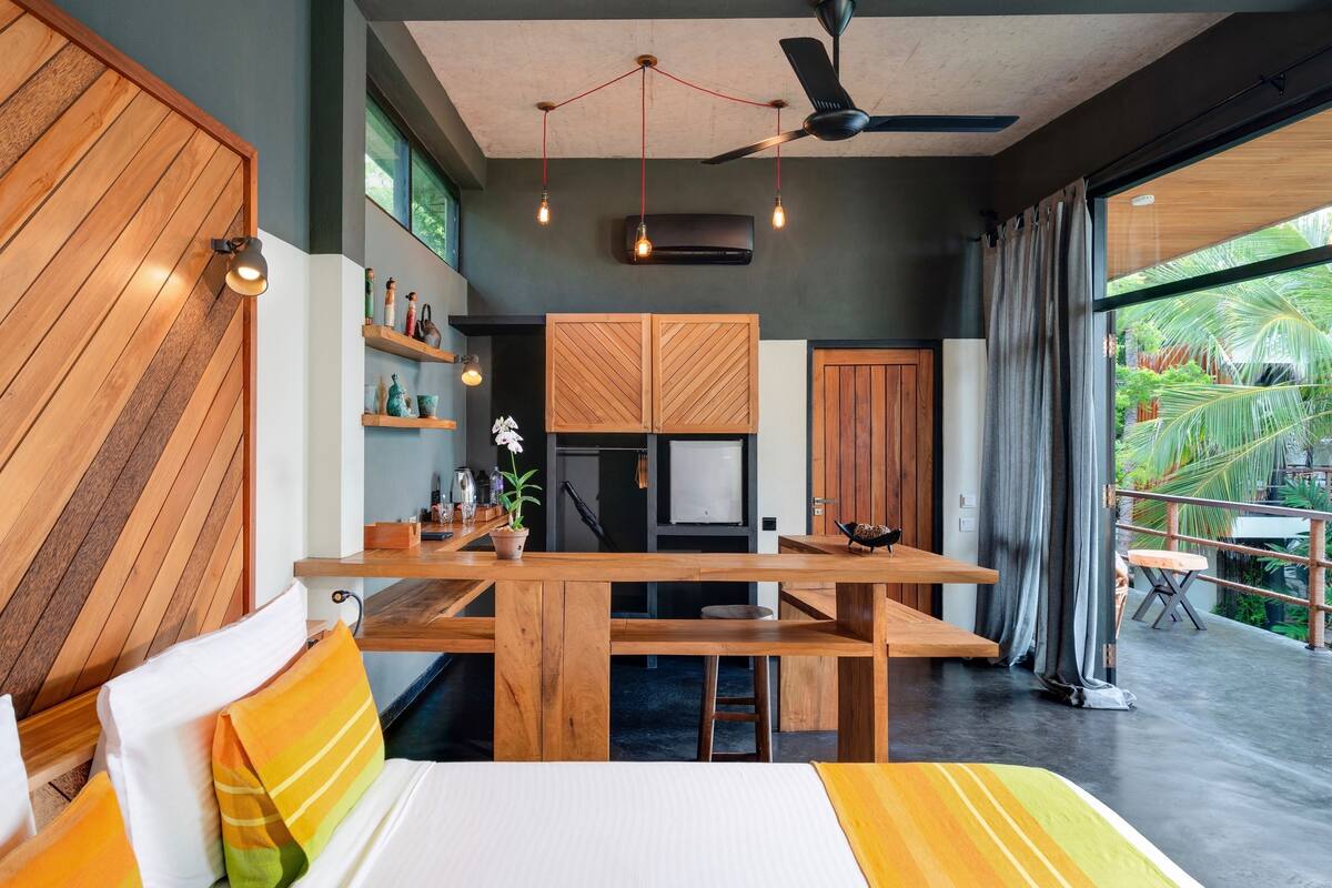A modern interior with a wooden dining table, a bed with colorful pillows, a ceiling fan, and a view of greenery outside.