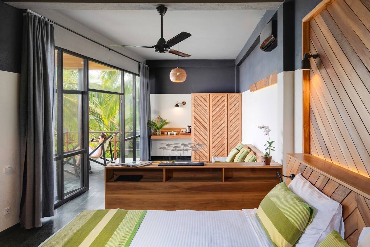 A modern bedroom with a large bed, a wooden headboard, a desk with a chair, and a view of palm trees through the window.
