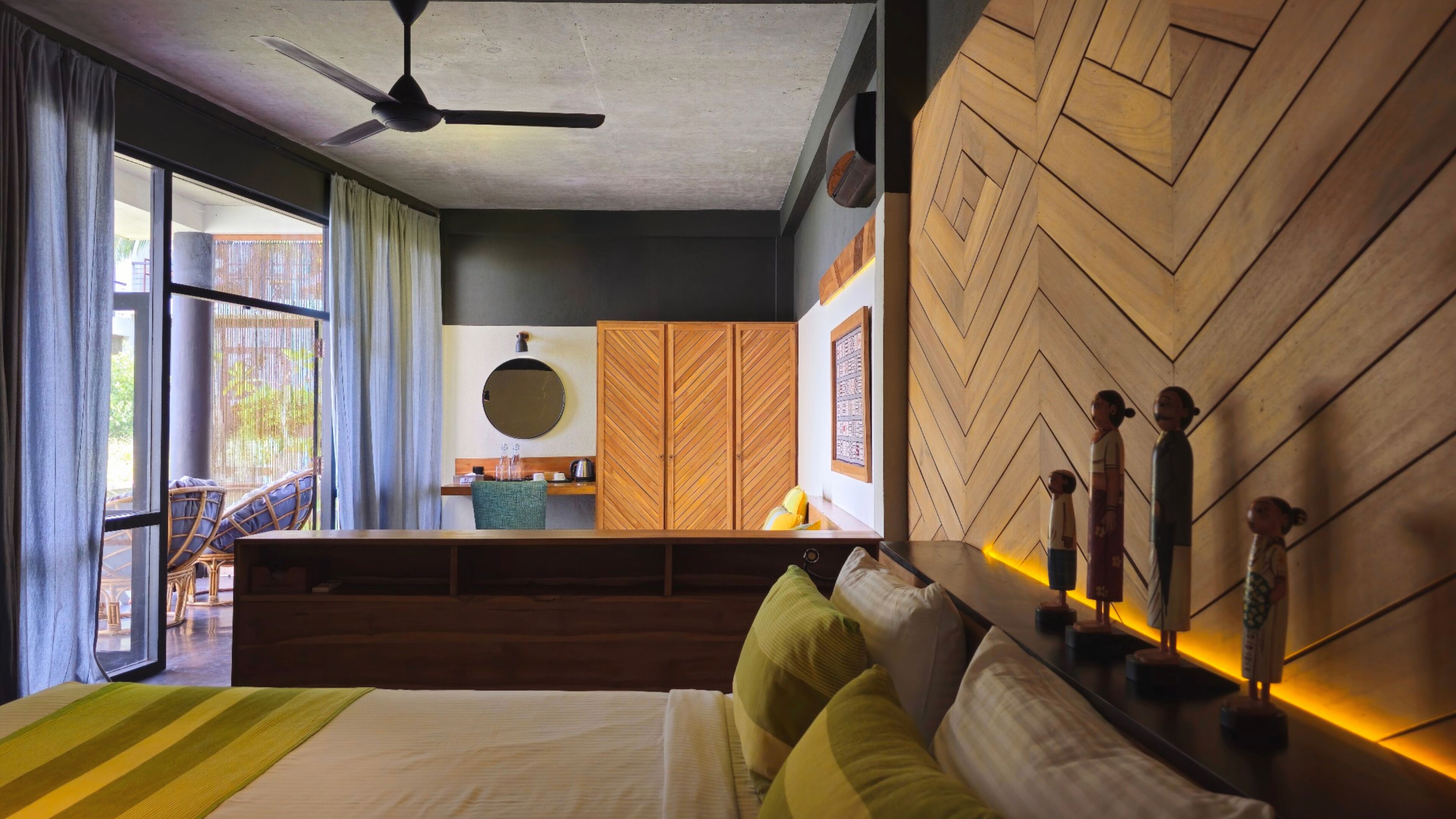 A hotel room with a bed, a desk, a chair, a ceiling fan, and a view of the outside.