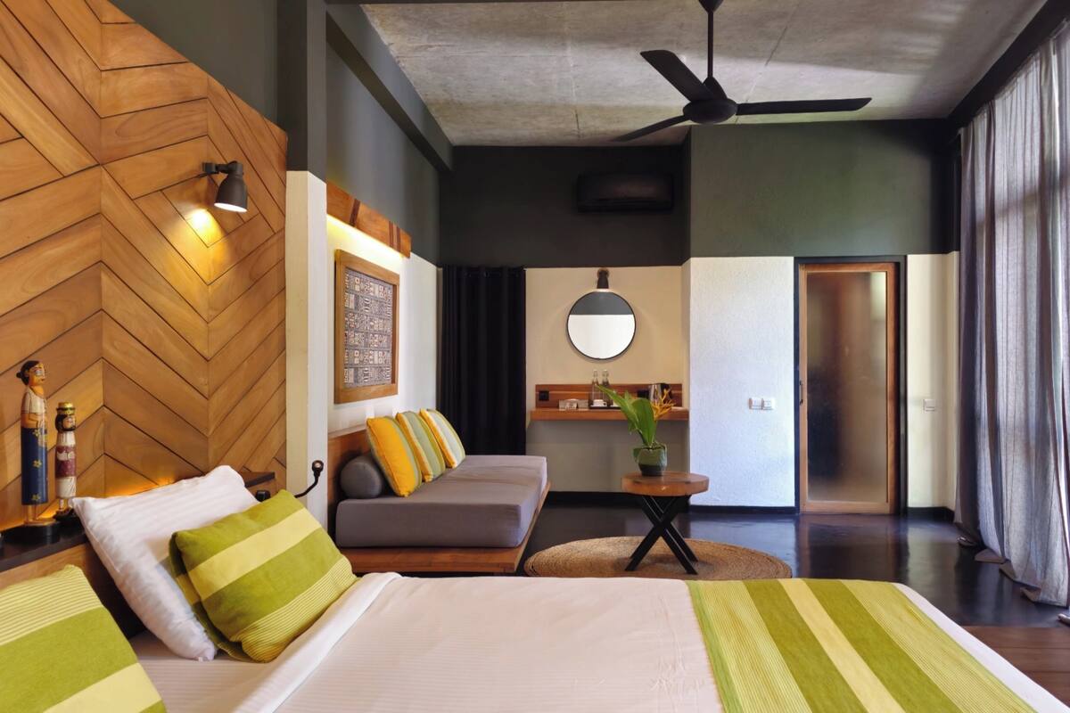 A hotel room with a wooden herringbone wall, a bed with green striped bedding, a gray sofa, a round mirror, and a ceiling fan.