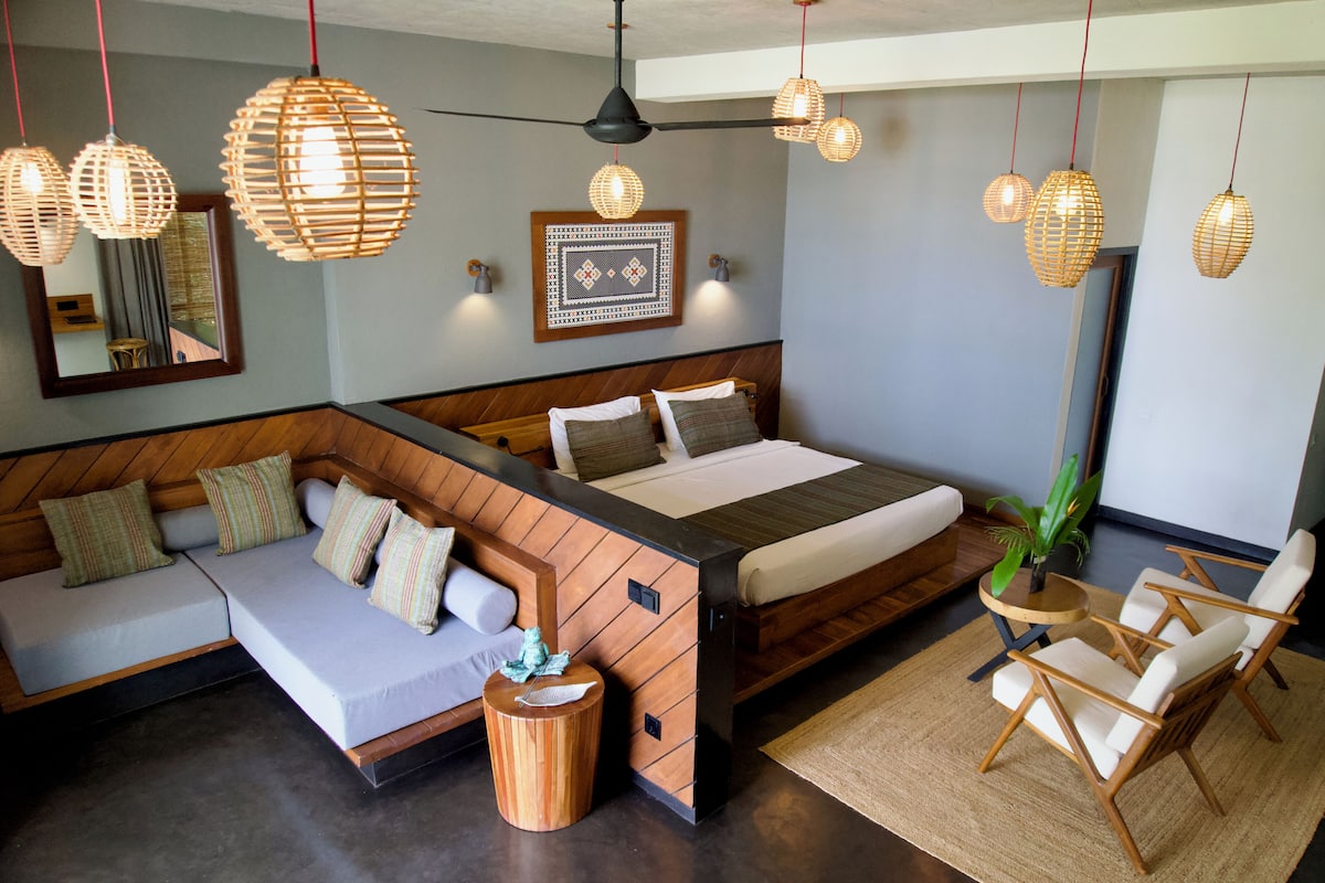 A modern bedroom with a large bed, a sofa, and two chairs. There are hanging lights and a mirror on the wall.