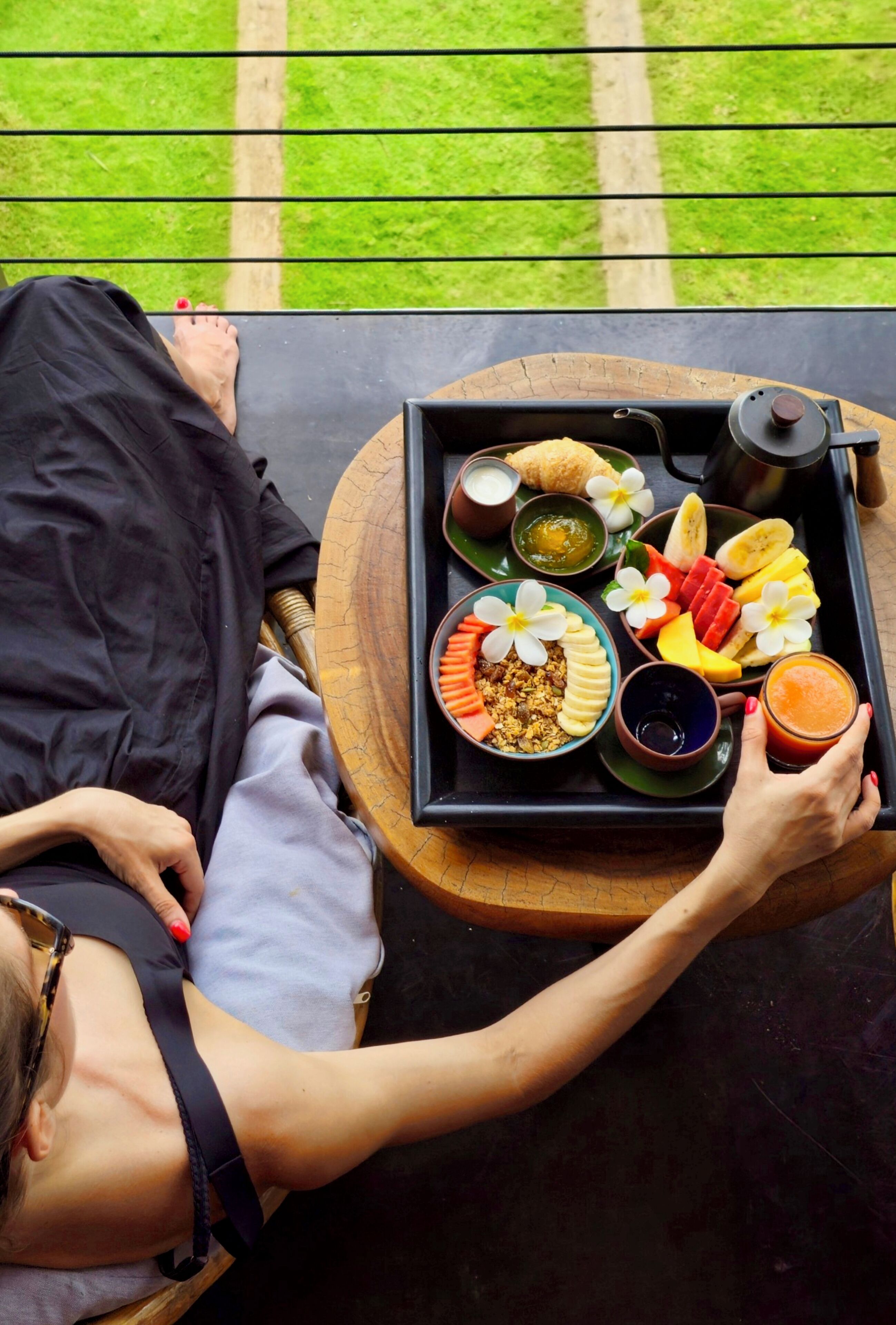 A tray with breakfast items including fruits, a drink, and a small bowl of yogurt, placed on a table outdoors.