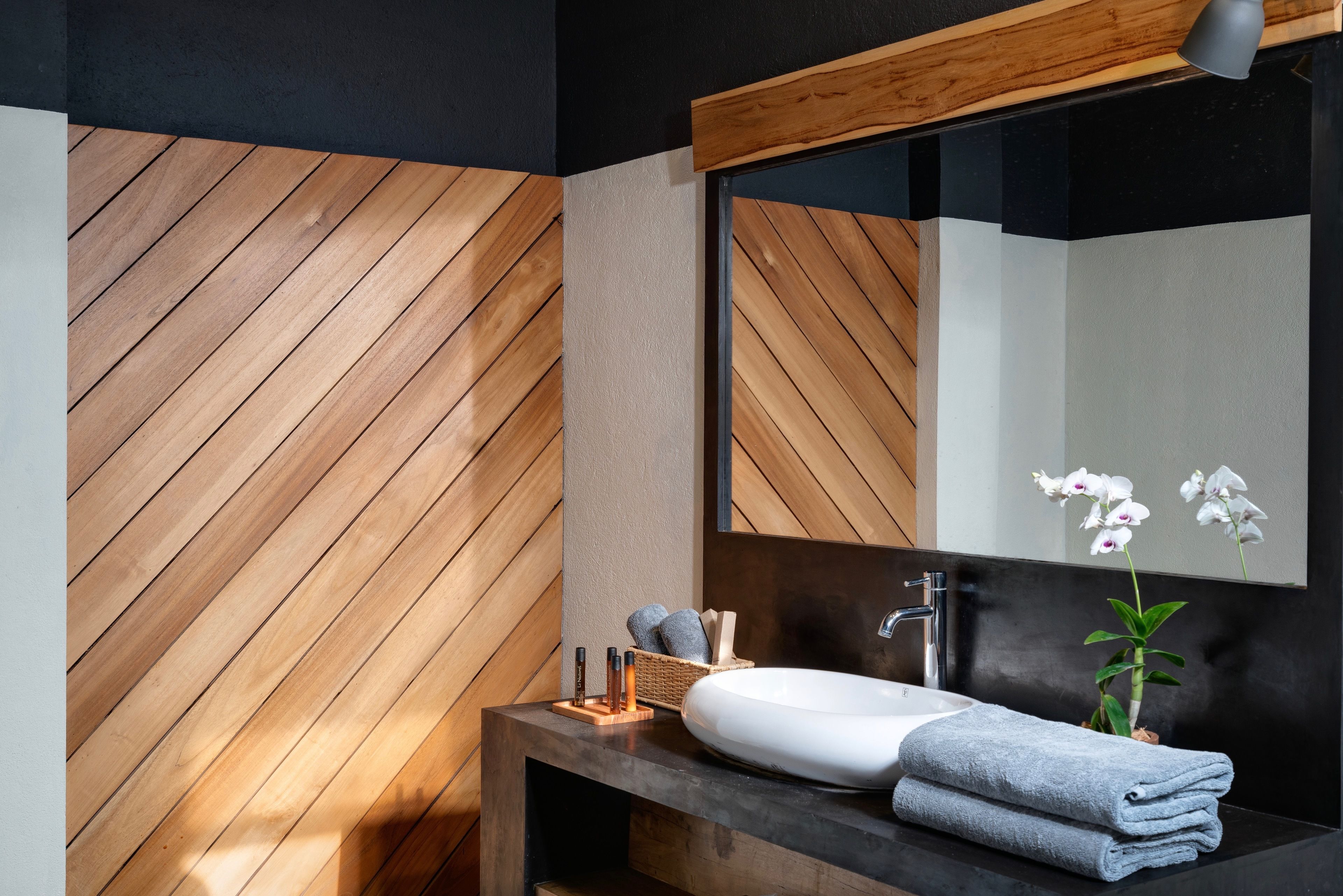 Um banheiro moderno com parede de madeira em padrão herringbone, um espelho grande, uma pia branca e uma pequena planta.