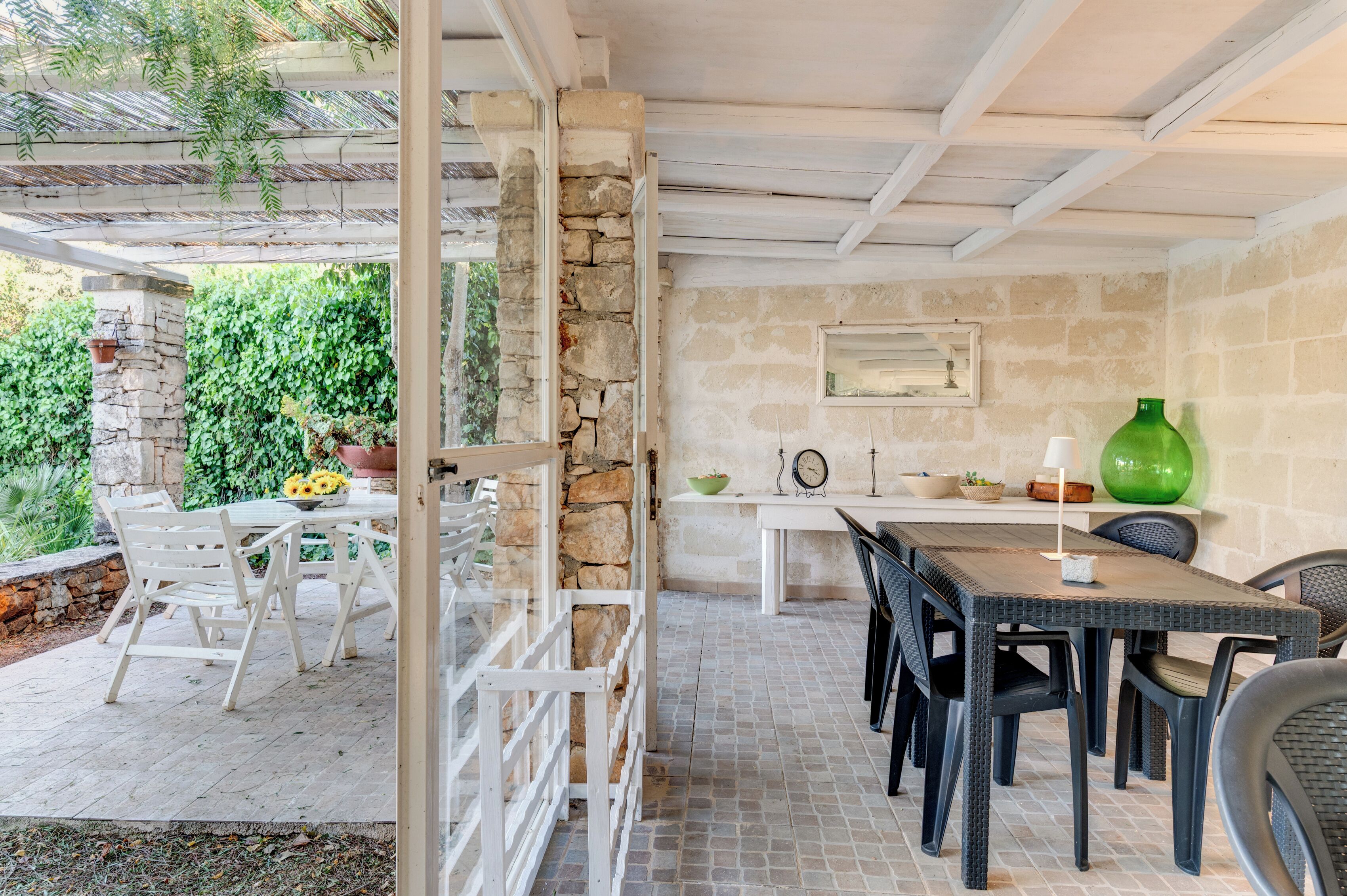 Ein möblierter Patio-Bereich im Freien mit einer Steinsäule, einem weißen Tisch mit Stühlen und einer grünen Vase auf einem Regal.
