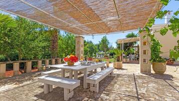 An outdoor patio with wooden tables and benches, surrounded by potted plants and a pergola.