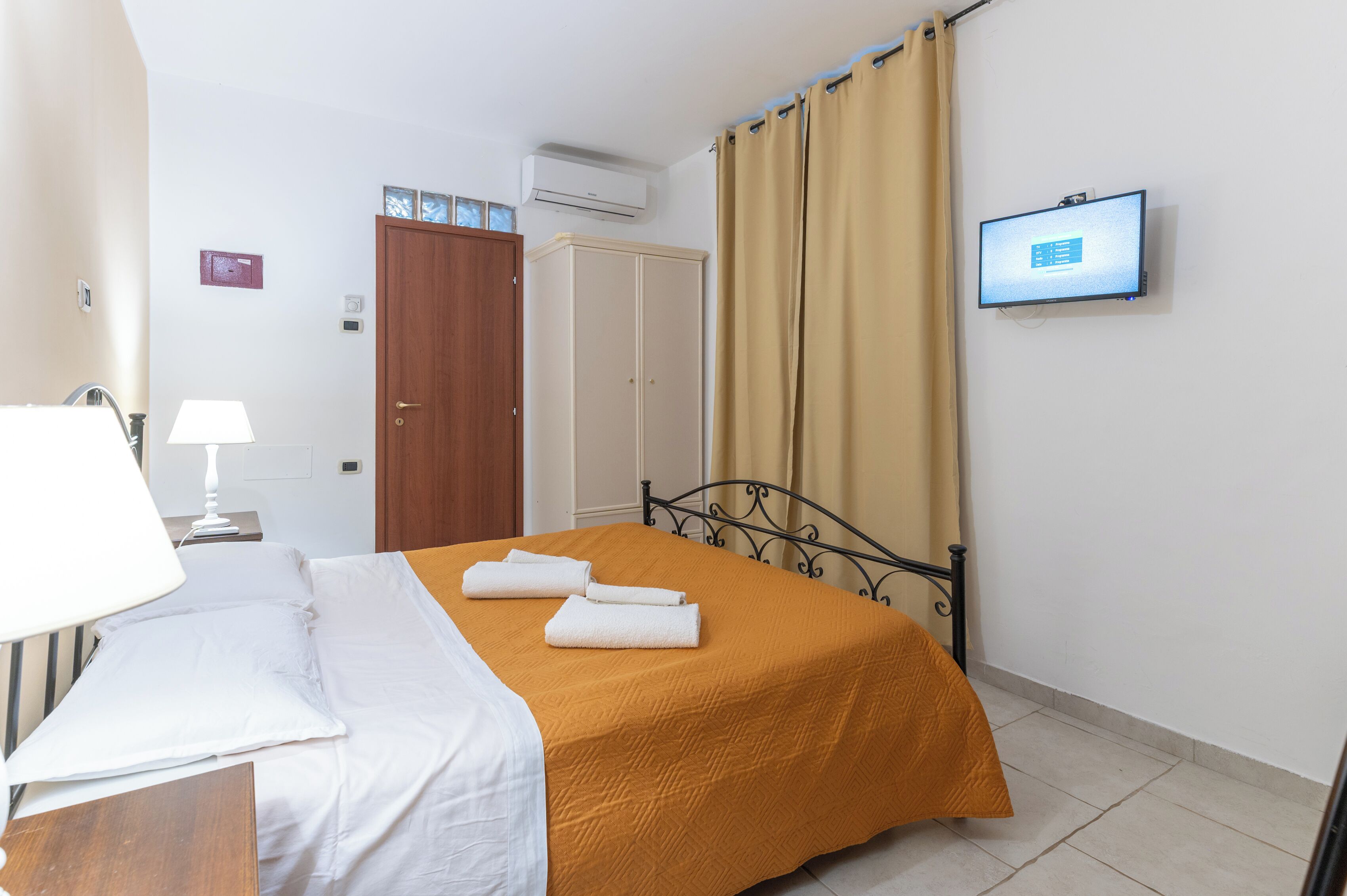A hotel room with a bed, a desk with a lamp, a wardrobe, a television on the wall, and a window with curtains.