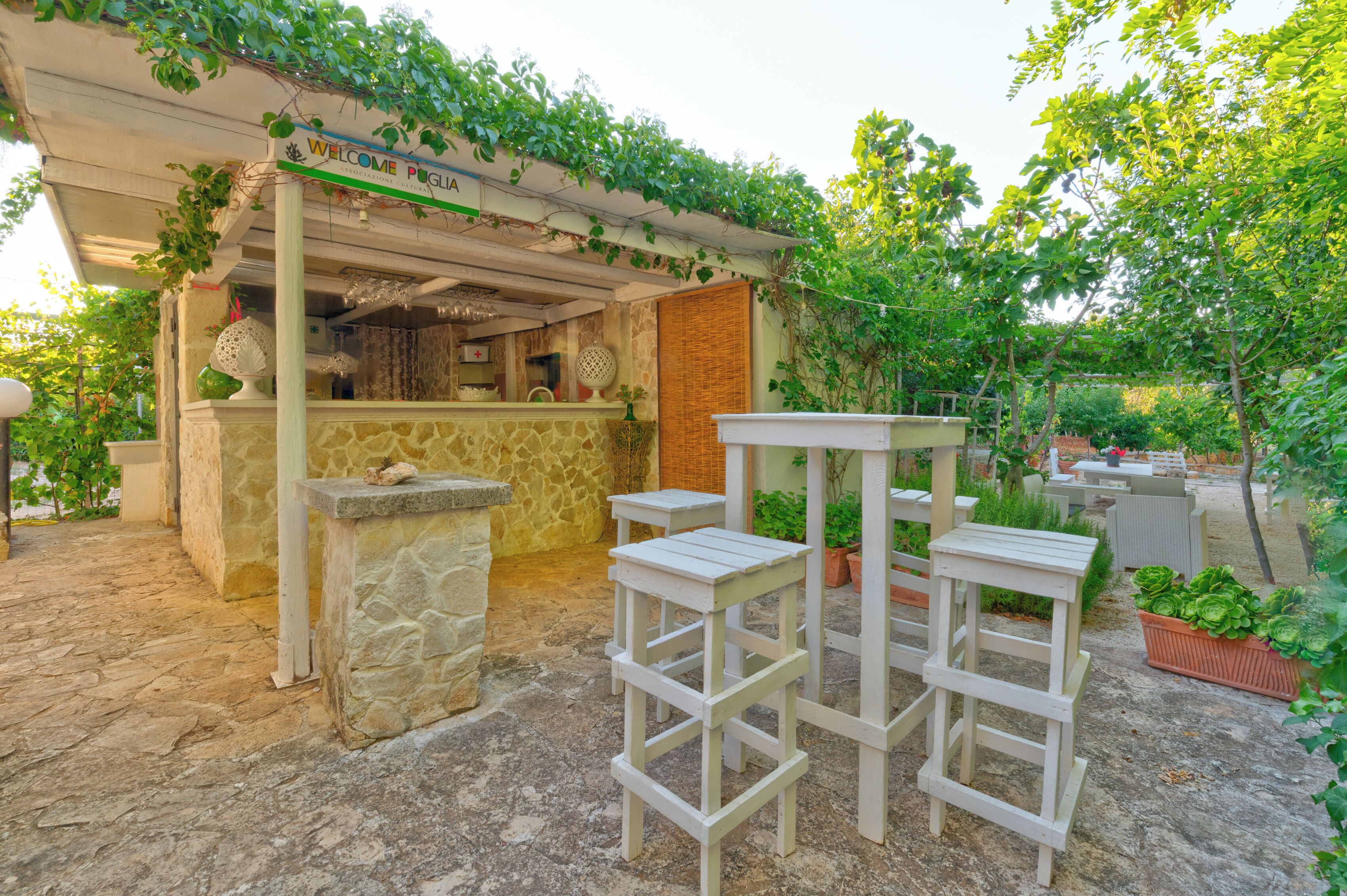 A patio area with a stone floor, white wooden tables and chairs, and a pergola with climbing plants.