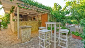 Un'area patio con pavimento in pietra, tavoli e sedie in legno bianco e un pergolato ricoperto di piante rampicanti.