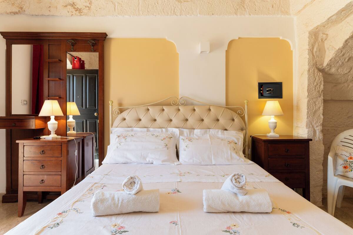 Un lit bien fait, avec deux serviettes, une table de chevet en bois avec une lampe, un miroir et un mur en pierre.