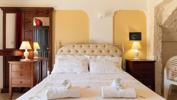 A neatly made bed with two towels, a wooden nightstand with a lamp, a mirror, and a stone wall.