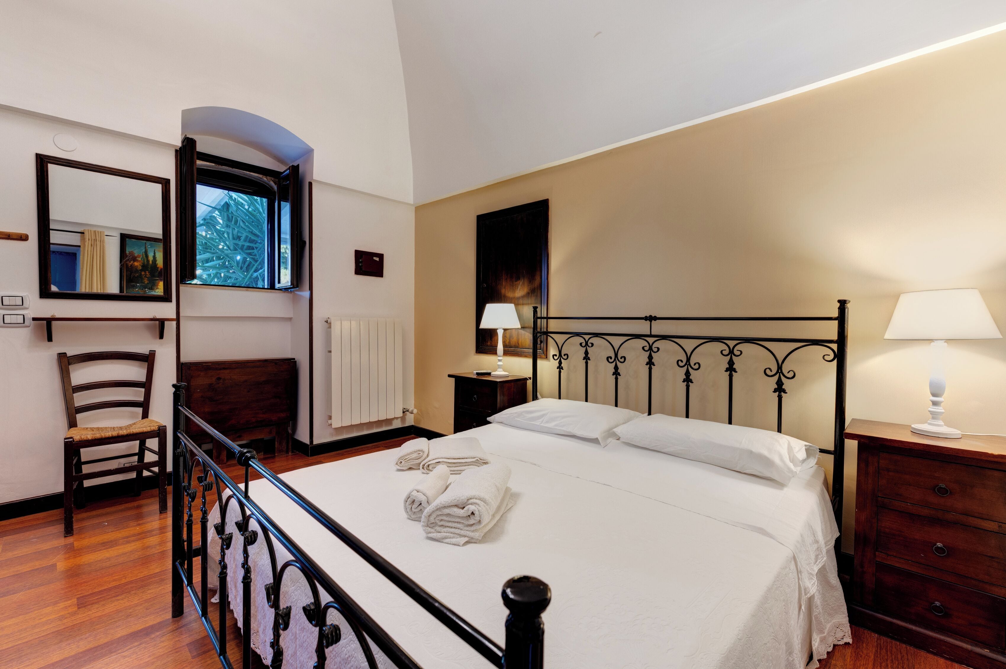 Cama bem arrumada com lençóis brancos, uma cadeira de madeira, mesa de cabeceira com abajur e janela com vista para árvores.