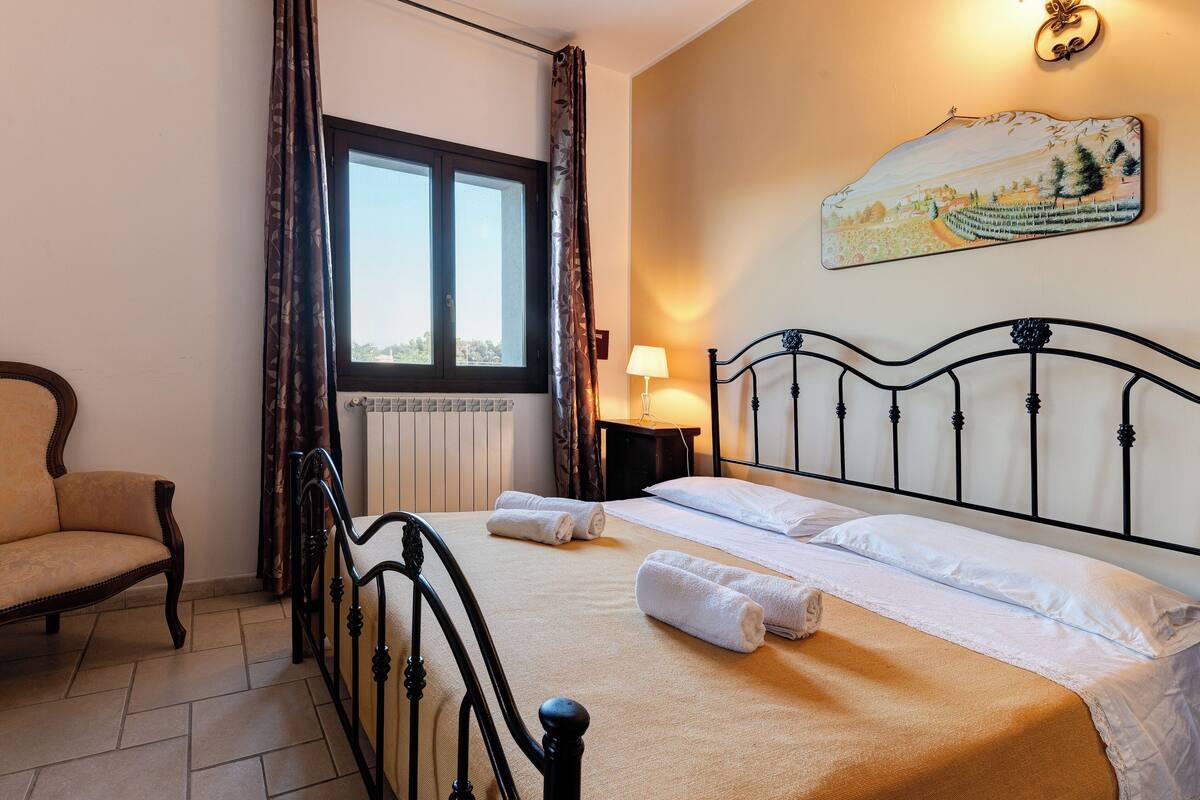 Un lit bien fait, avec du linge de lit et des serviettes blanches, une chaise en bois, une fenêtre avec des rideaux, une table de chevet avec une lampe et un tableau accroché au mur.