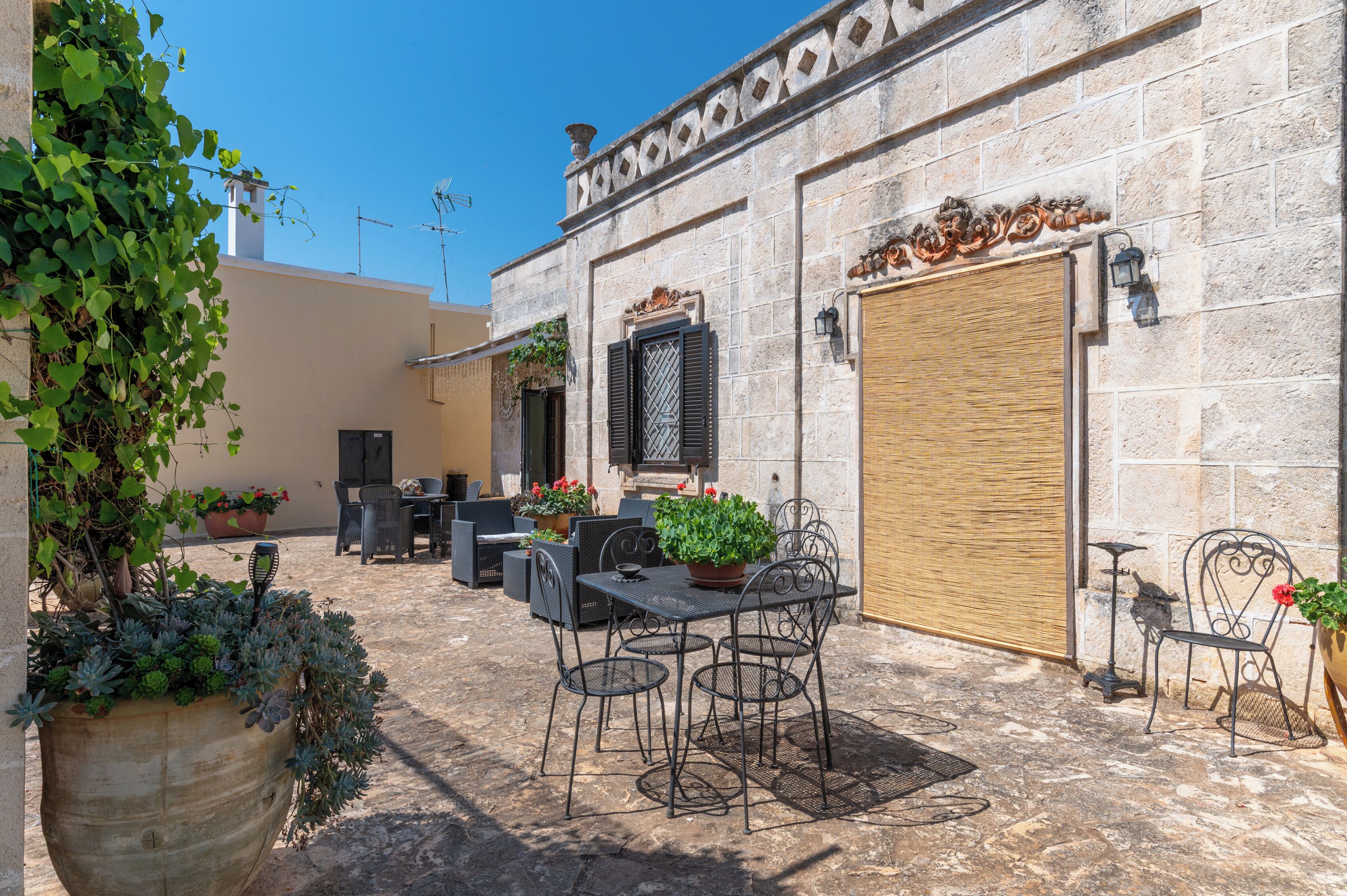 Une terrasse extérieure avec une table et des chaises, entourée de plantes en pot et d’un mur de pierre.