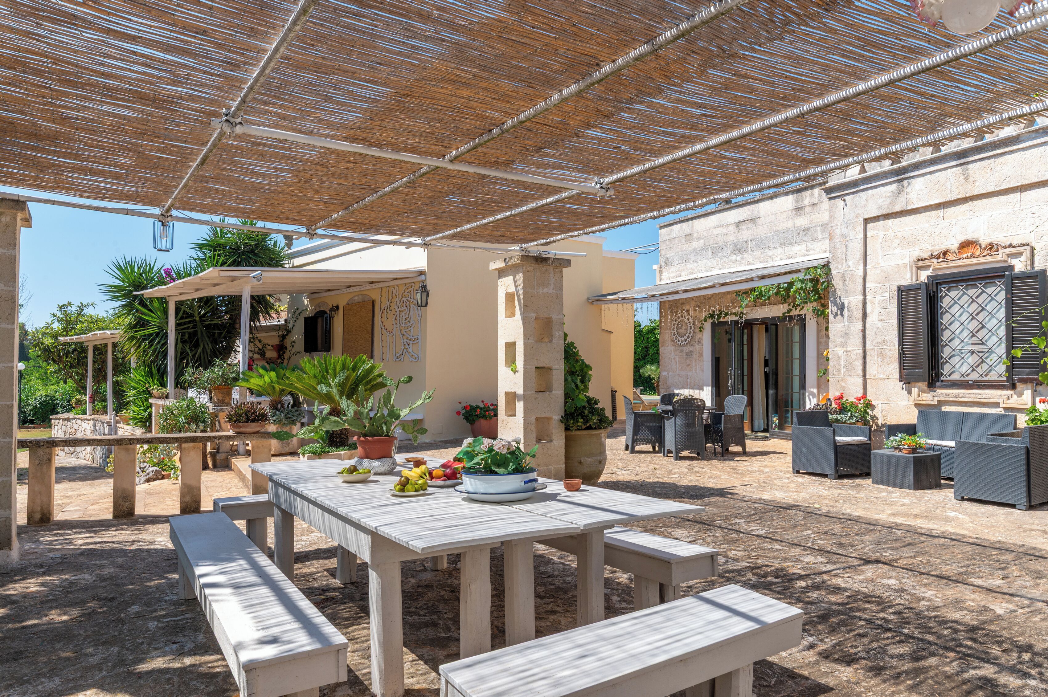 Een terras buiten met houten tafels en banken, omgeven door plantenpotten en een pergola.