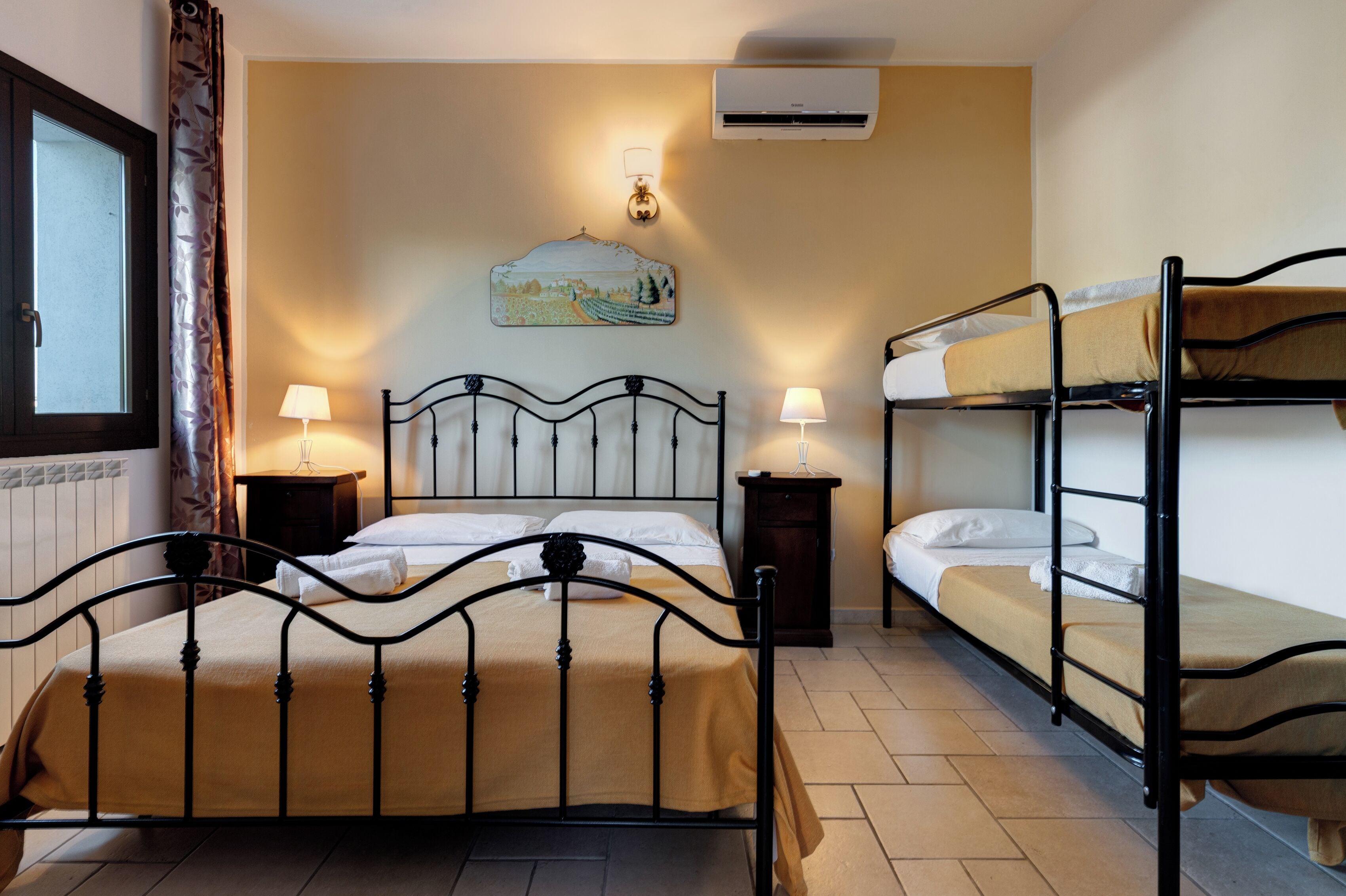 A hotel room with a single bed, two bedside tables, a bunk bed, an air conditioning unit, and a window with curtains.