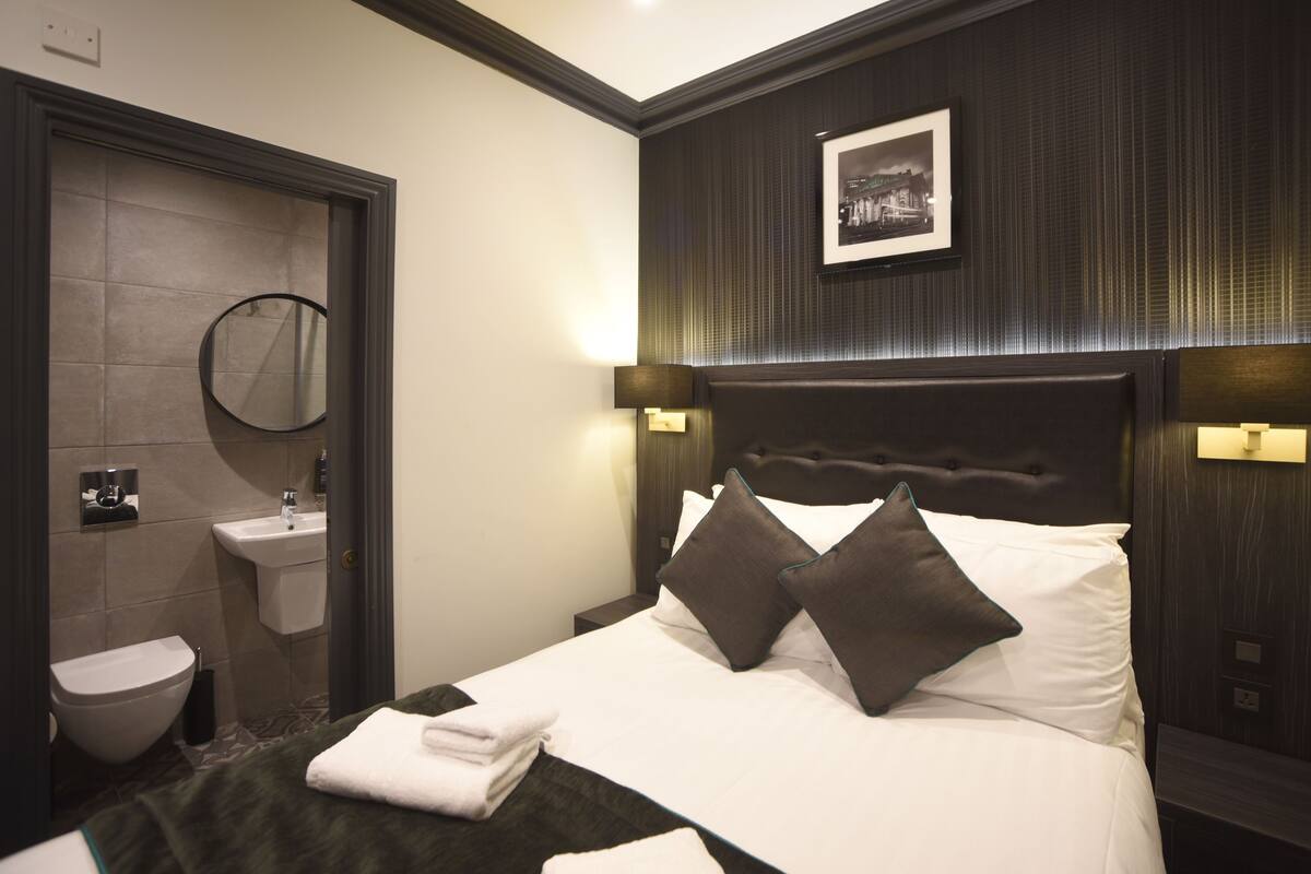 A hotel room with a bed, a bedside table, a wall-mounted picture, a bathroom with a sink and toilet, and a mirror.