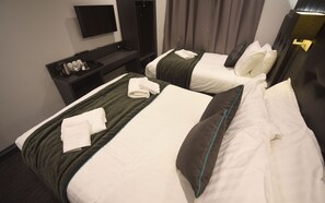 Duas camas bem arrumadas com roupa de cama branca e detalhes verdes, uma televisão de tela plana na parede, uma mesinha com artigos de higiene pessoal e um guarda-roupa de cor escura.