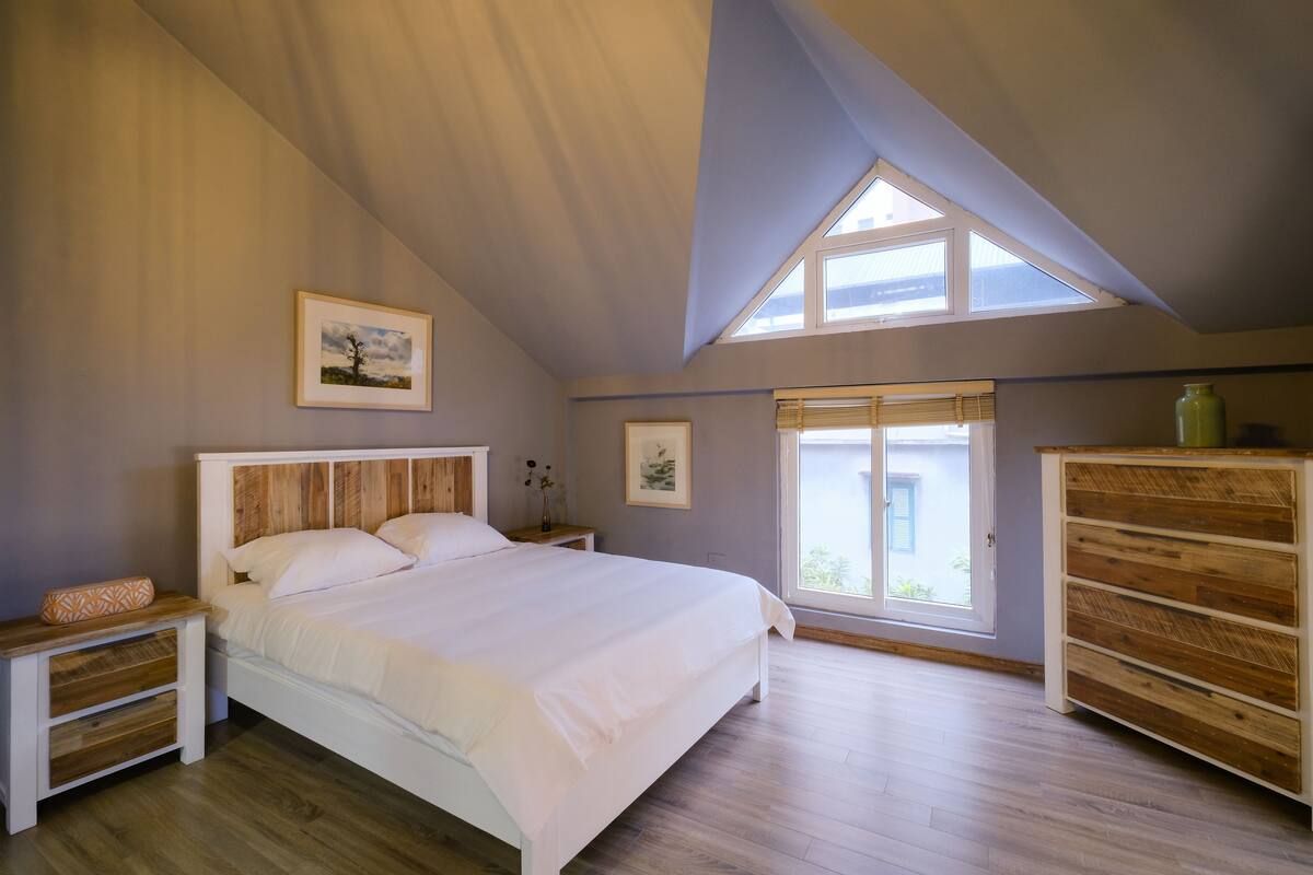 Un dormitorio con techo inclinado, una cama grande, mesitas de noche de madera, un aparador y dos ventanas que dejan entrar luz natural.