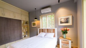 A bedroom with a bed, a nightstand, a window with blinds, a wall-mounted air conditioner, and a shelf with decorative items. - Le Petit Hanoi (Hanoi)