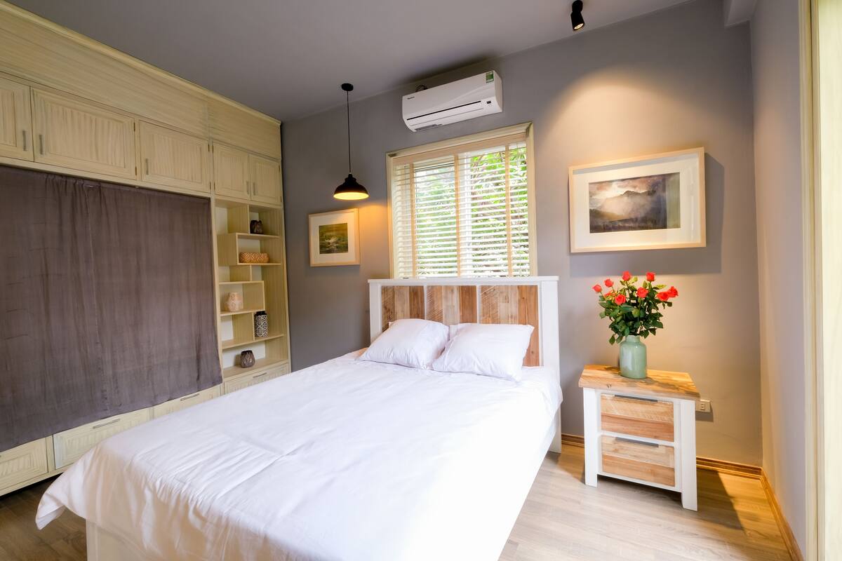 Un dormitorio con una cama, una mesita de noche, un ventanal con persianas, un aire acondicionado de pared y una estantería con objetos decorativos.