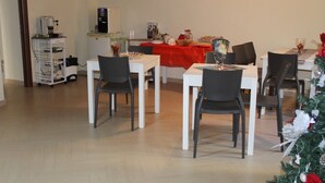 Un comedor con mesas blancas y sillas negras, una cafetera y un refrigerador.
