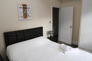 A neatly made bed with white linens and a rolled towel, a black upholstered headboard, a wall-mounted artwork, and a white door in a room with beige walls. - Guards View Windsor (Windsor)
