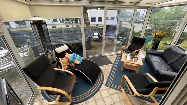 Ein sonnendurchfluteter Balkon mit einer schwarzen Ledersofa, zwei Holzstühlen, einem kleinen Tisch und einem Grill.