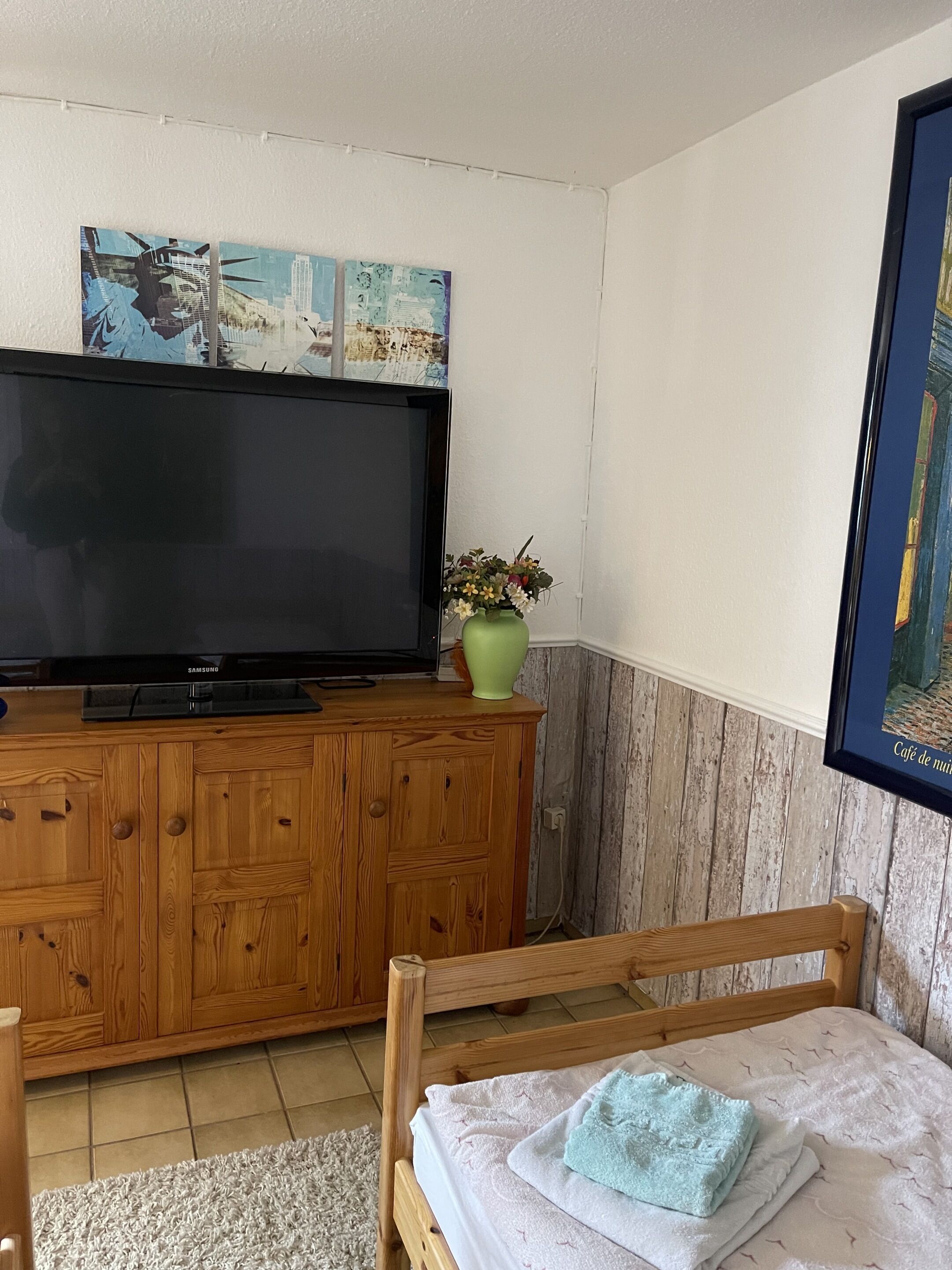 Ein Schlafzimmer mit einem Bett, einem Holzschrank mit Fernseher, einem grünen Blumenvasen und einem gerahmten Bild an der Wand.