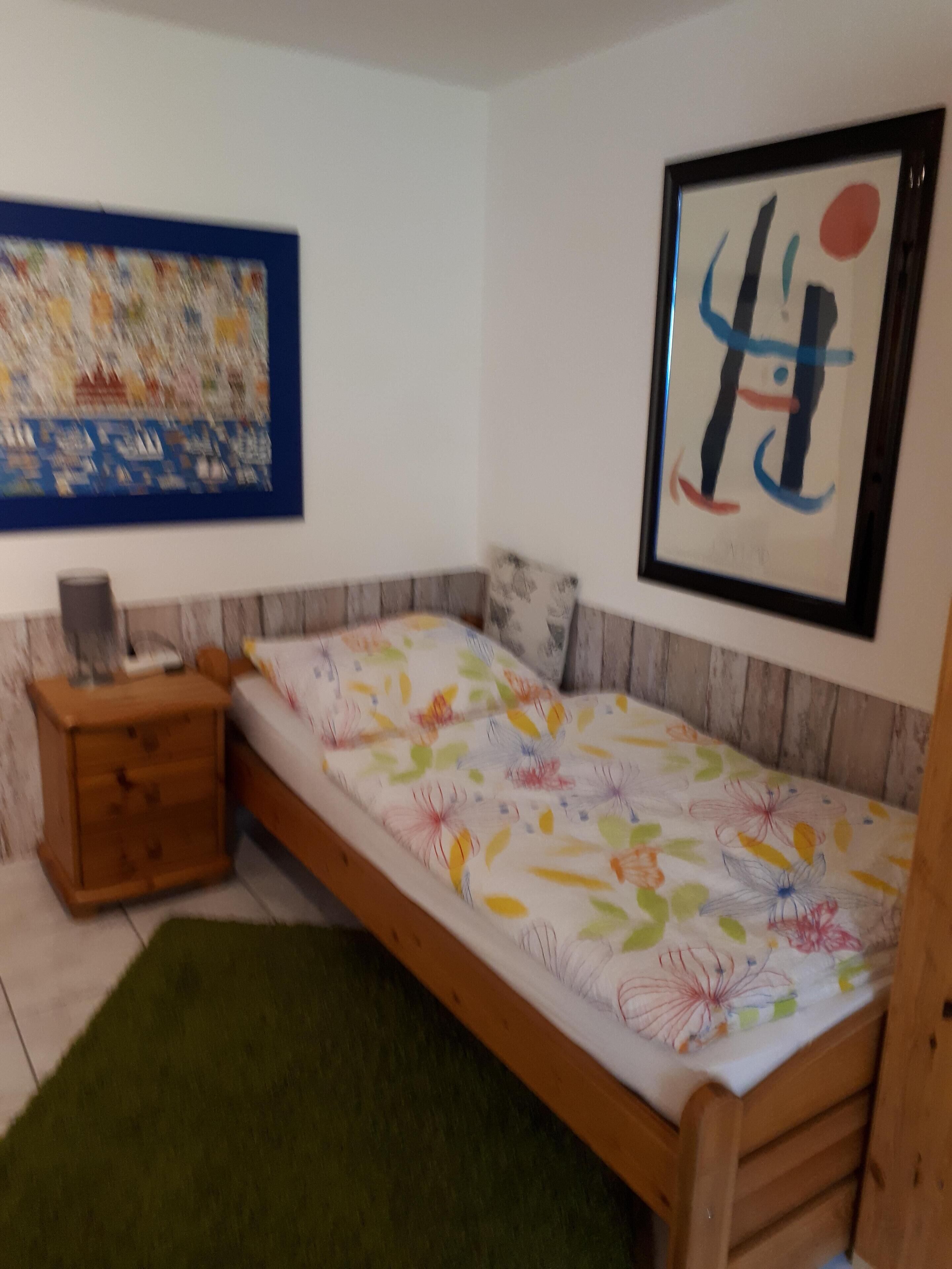 Pieni huone, jossa on yksi sänky, puinen pääty, vihreä matto, pieni puinen yöpöytä, työpöytä ja kaksi kehystettyä taideteosta seinillä.