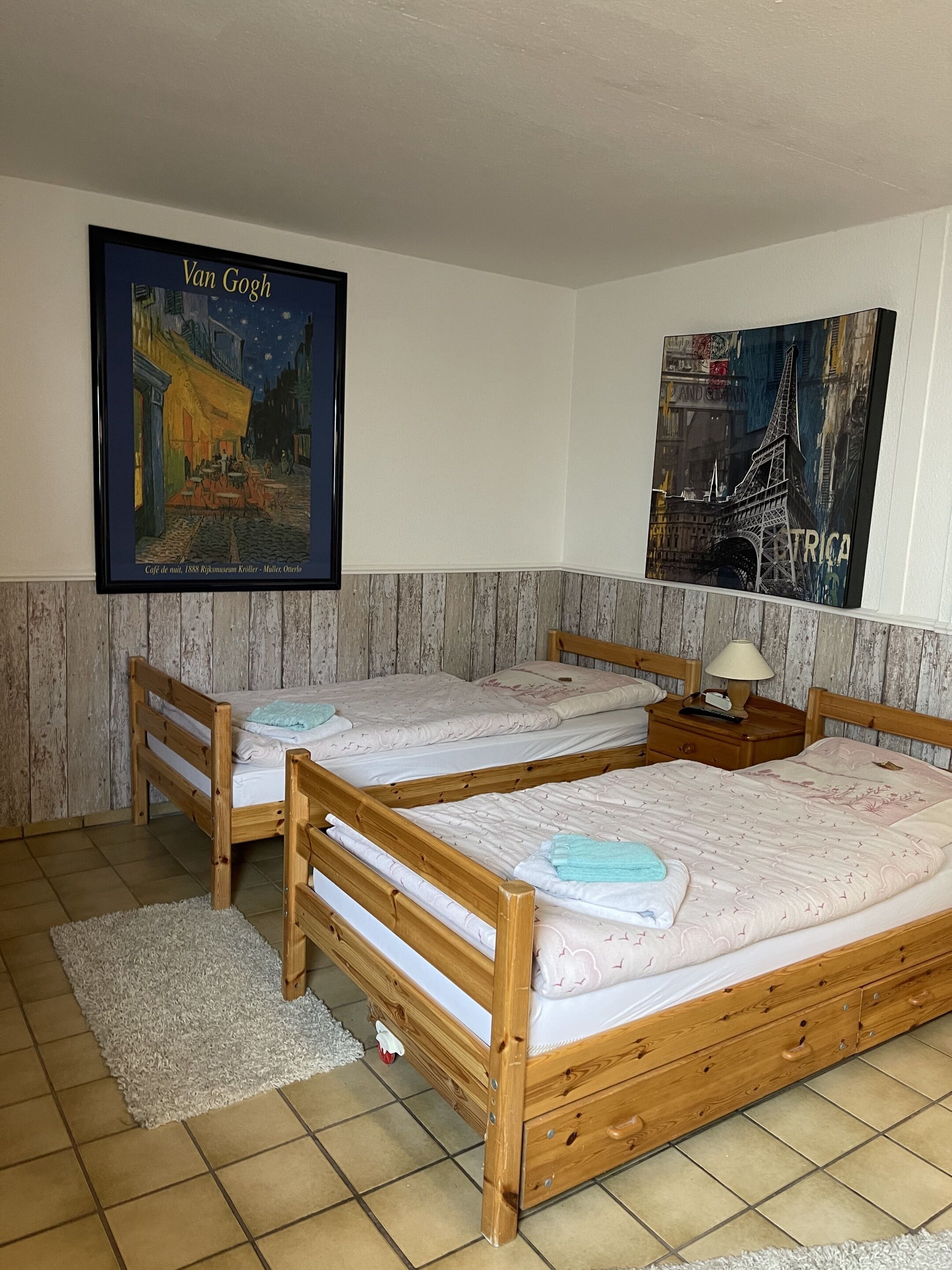 A room with two wooden beds, a nightstand with a lamp, and two framed artworks on the wall.