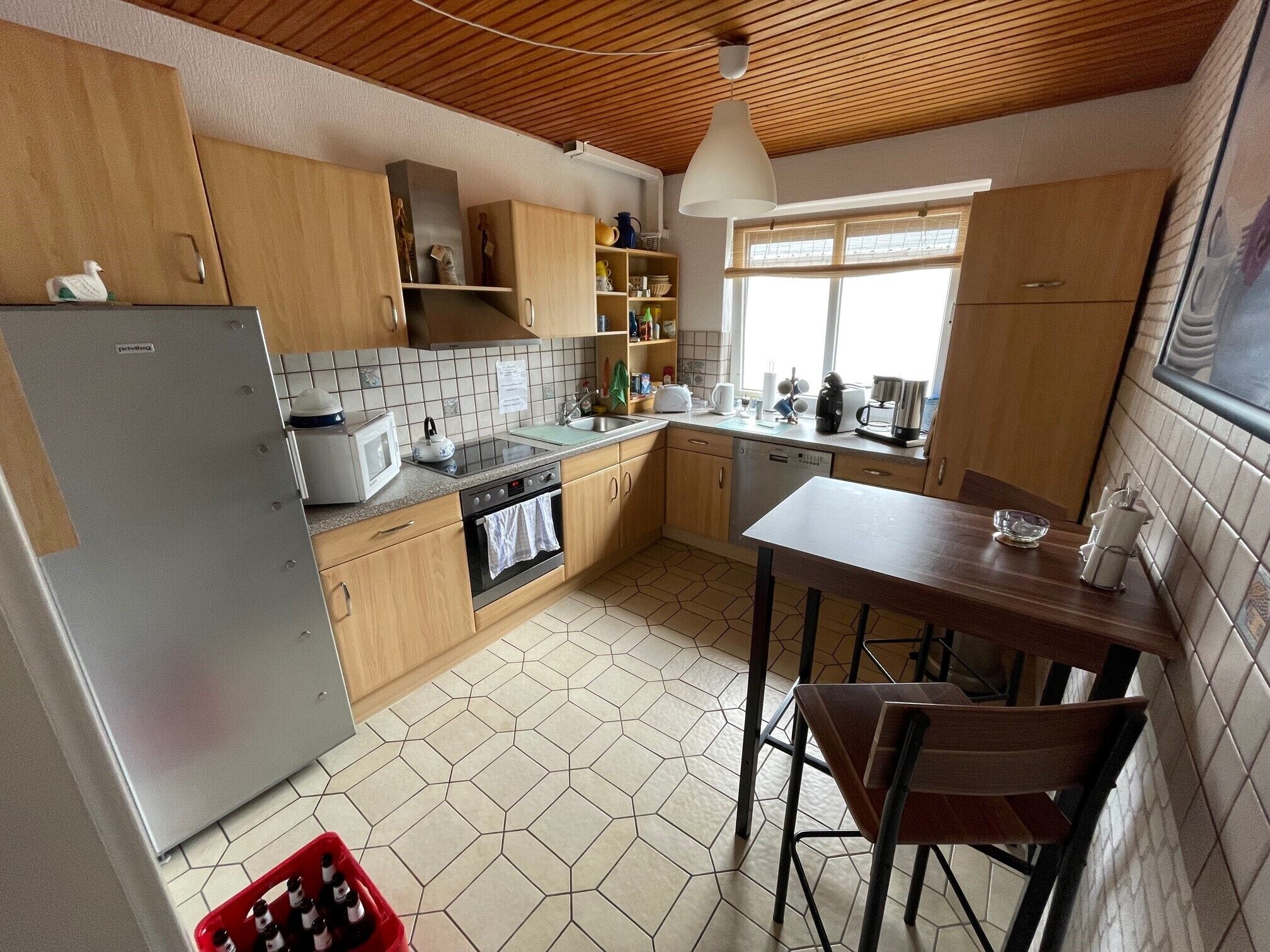 A kitchen with wooden cabinets, a stainless steel refrigerator, a tiled backsplash, a window with blinds, and a dining area with a table and chairs.