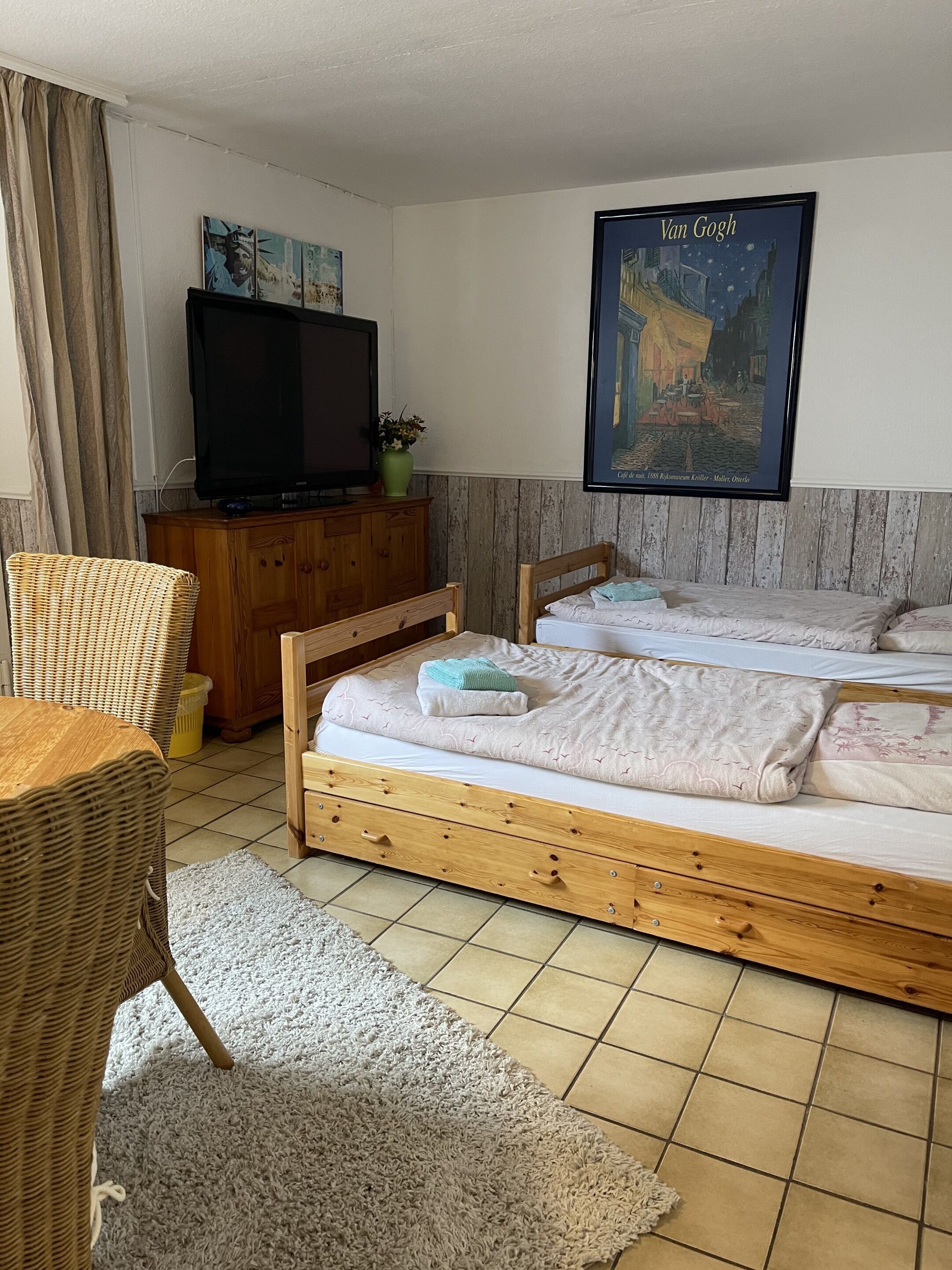 Ein Schlafzimmer mit zwei Holzbetten, einem Fernseher auf einem Holzschrank, einem Korbstuhl und einem Bild von Van Goghs 'Café Terrasse am Abend' an der Wand.