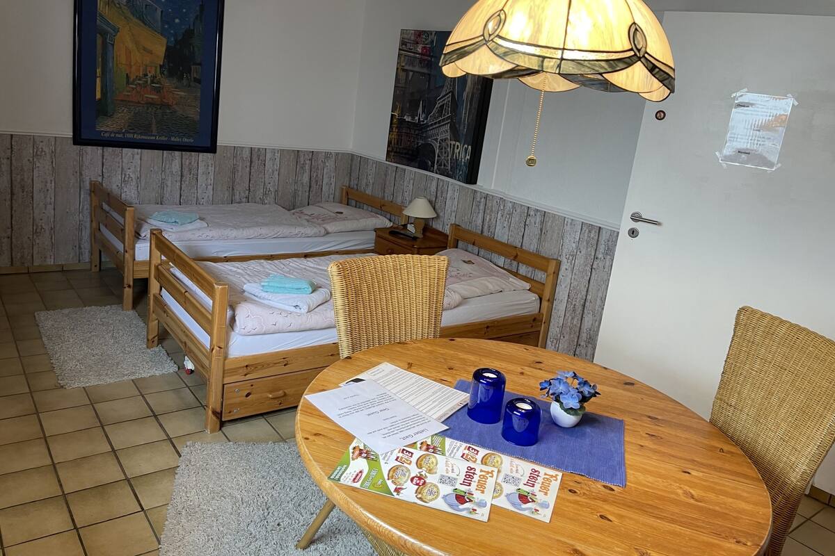 Ein Zimmer mit zwei Holzbetten, einem Esstisch mit Papieren und blauen Kerzen sowie einer Hängelampe.
