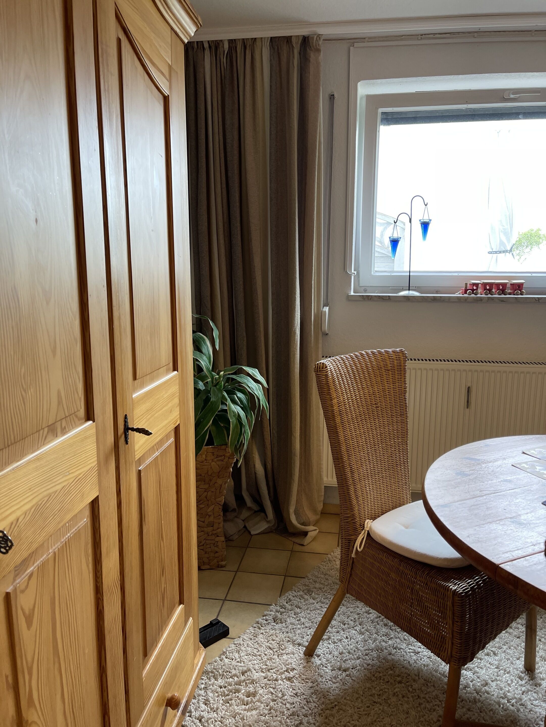 A room with a wooden wardrobe, a wicker chair, a round table, a potted plant, and a window with curtains.