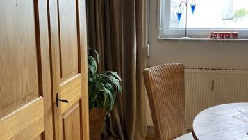 A room with a wooden wardrobe, a wicker chair, a round table, a potted plant, and a window with curtains.