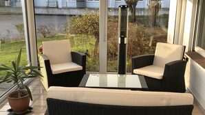 Eine Terrassenfläche mit zwei schwarzen Korbstühlen, weißen Kissen, einem Glastisch und einem Blumentopf.