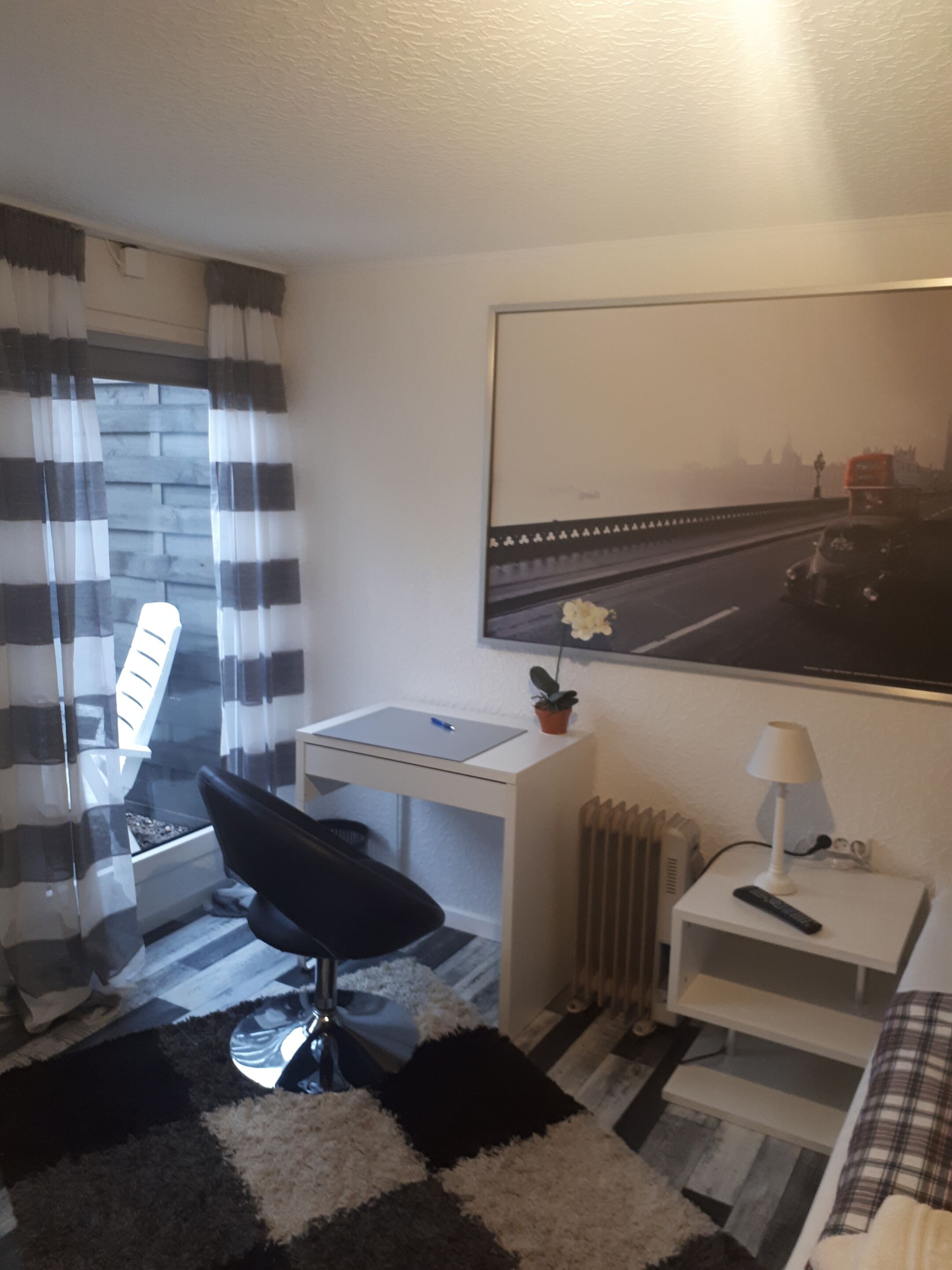 A compact hotel room with a bed, a desk, a chair, a radiator, and a window with curtains.