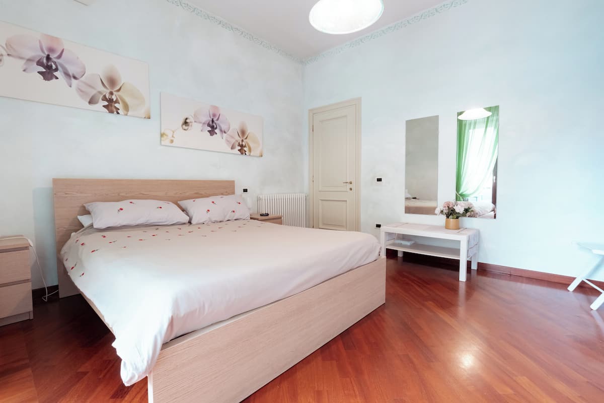 A bedroom with a large bed, a bedside table, a nightstand, a dresser, a mirror, and a window with curtains.