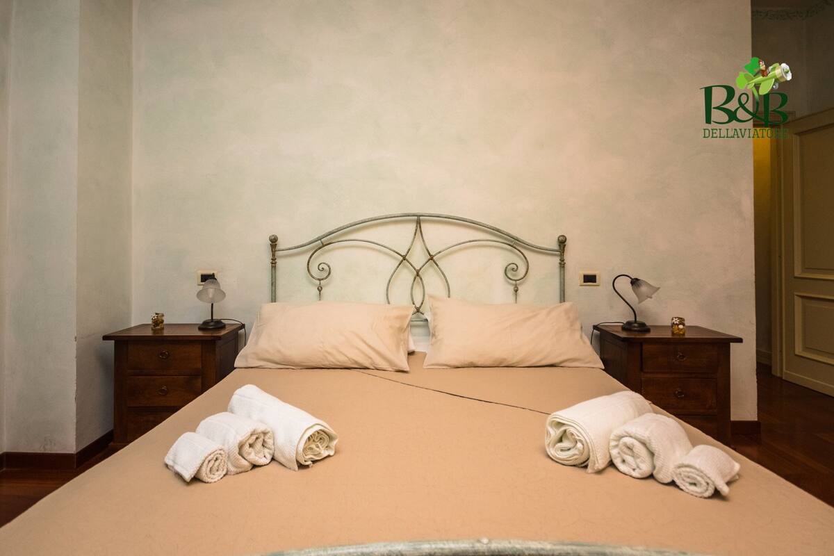 A neatly made bed with two pillows, two bedside tables with lamps, and rolled towels on the bed.