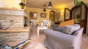 Un salon chaleureux avec une cheminée en pierre, un canapé gris et un coin repas avec des chaises blanches.