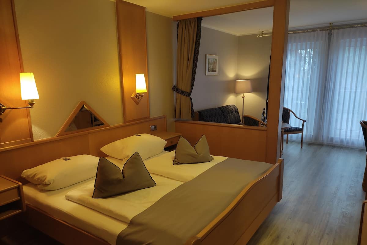 Une chambre d’hôtel avec un grand lit, des lampes de chevet, un miroir, une chaise et une vue sur l’extérieur à travers des rideaux légers.