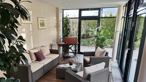 A modern living room with wicker furniture, a coffee table with papers, and a view of greenery outside. - Hotel Zum Märchenwald (Lingen)
