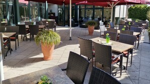 An outdoor patio with wicker tables and chairs, red umbrellas, and potted plants. - Hotel Zum Märchenwald (Lingen)