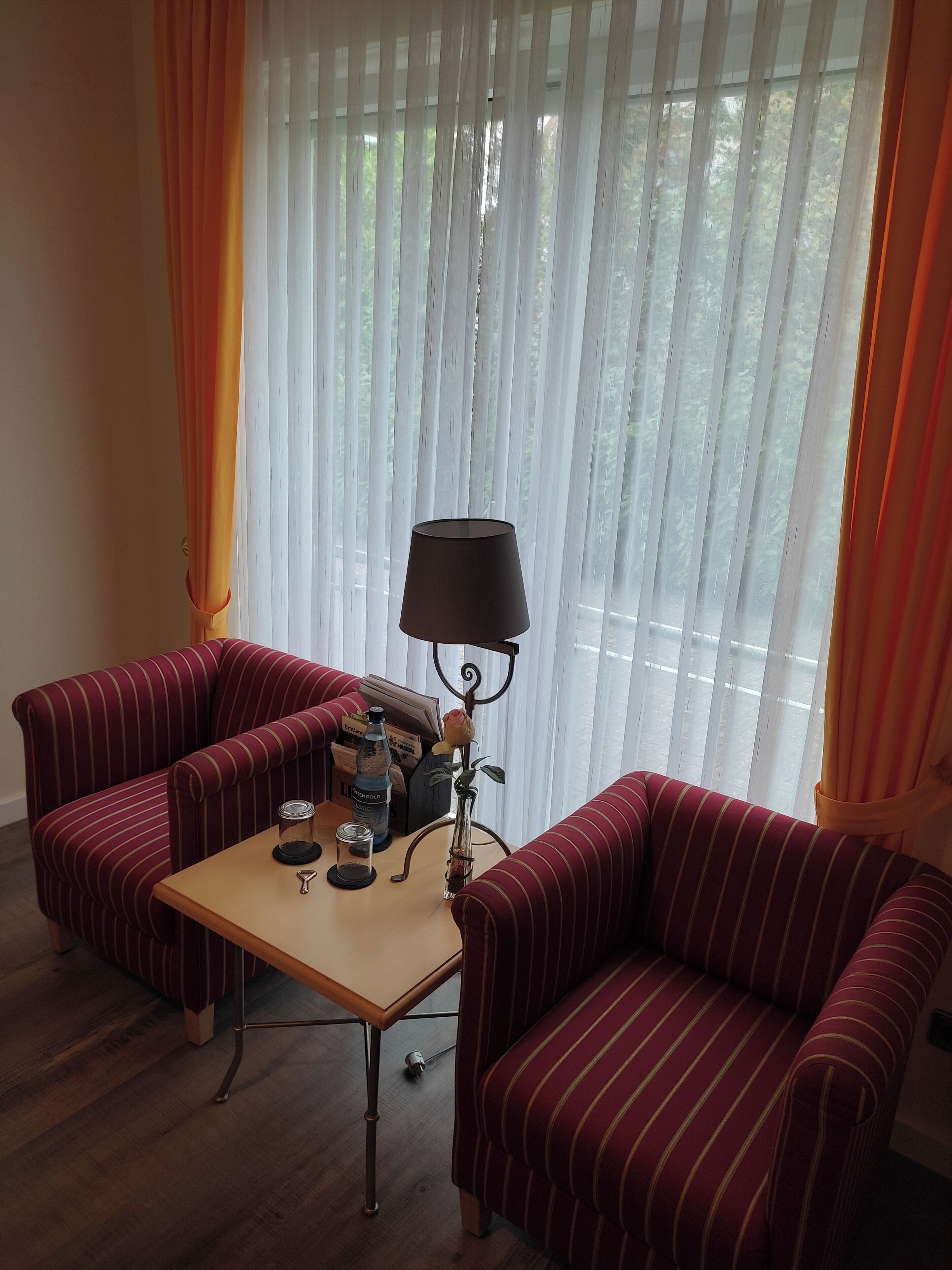Ein Zimmer mit zwei gestreiften Sesseln, einem kleinen Tisch mit Lampe und einem Fenster mit transparenten Vorhängen.